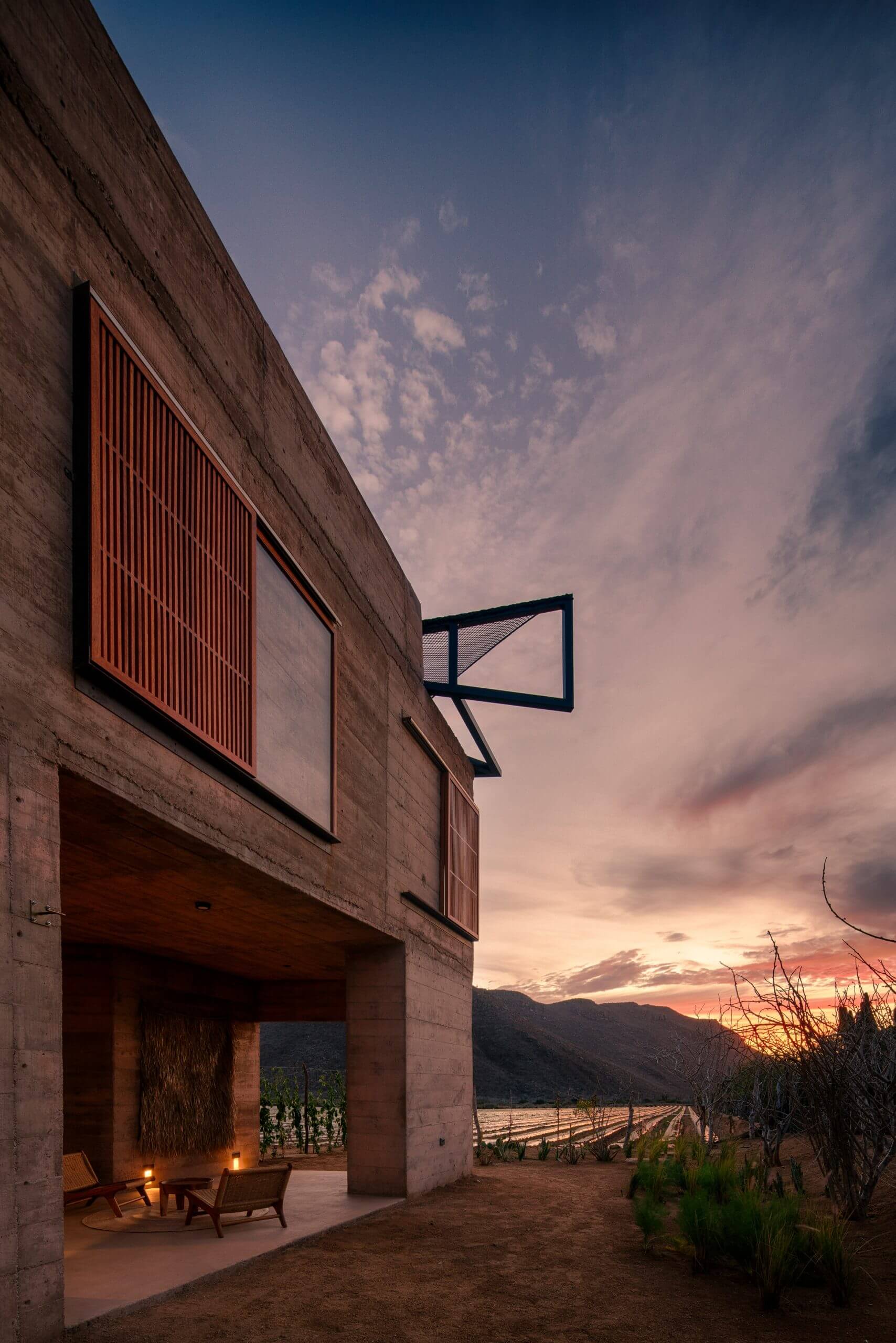Paradero Todos Santos, Mexico — Rammed earth façade with steel-framed windows overlooking vineyard valley at dusk, modernist hospitality architecture.