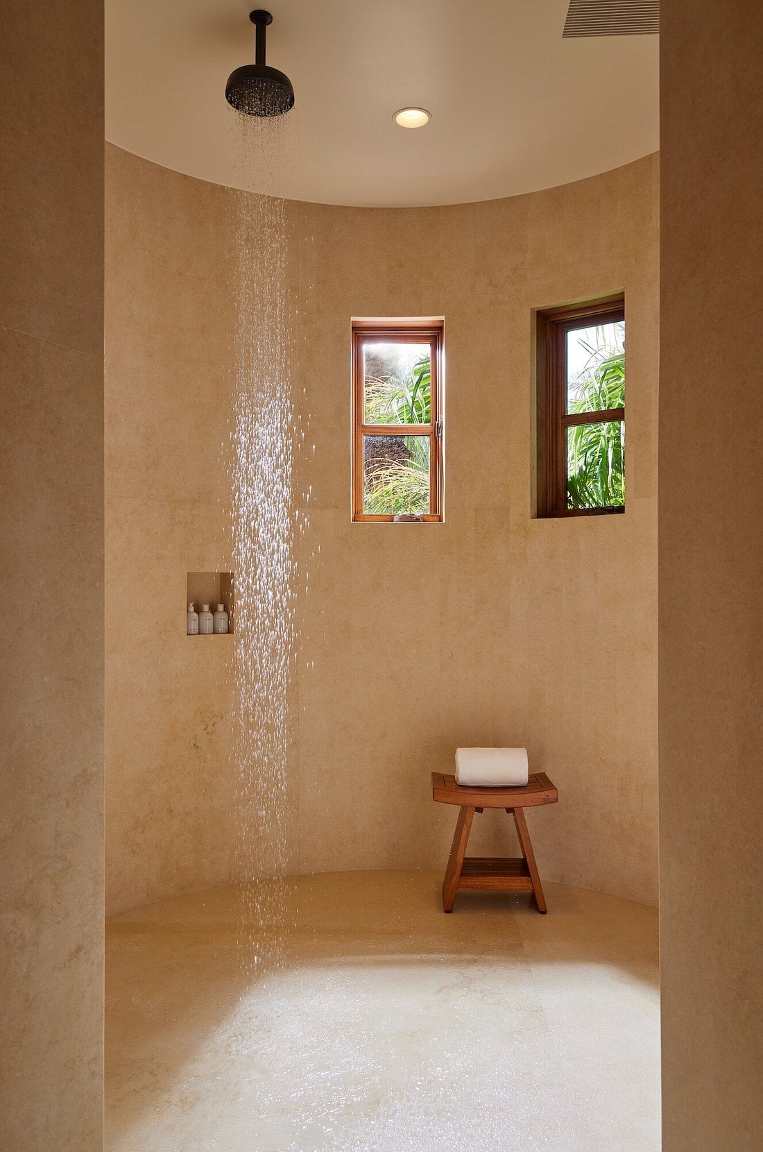 Sensei Porcupine Creek, North America — Luxury shower with curved travertine walls, rainfall showerhead, natural light from wooden-framed windows, and minimalist wooden stool.