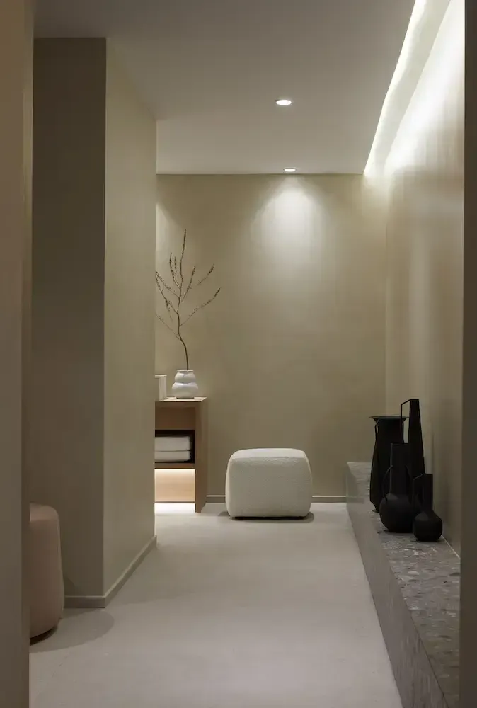 Pārocks, Europe — Minimalist entryway with soft ambient lighting, neutral stone flooring, white sculptural pouf, wooden shelving, and dried botanicals in ceramic vessel.