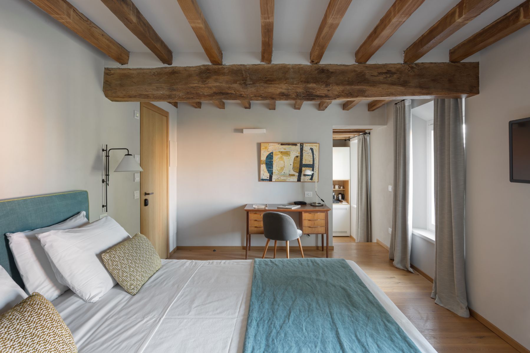 Roncolo 1888, Europe — Bedroom with exposed wooden beam ceiling, teal upholstered headboard, mid-century desk, warm wood floors, and minimalist modern furnishings.