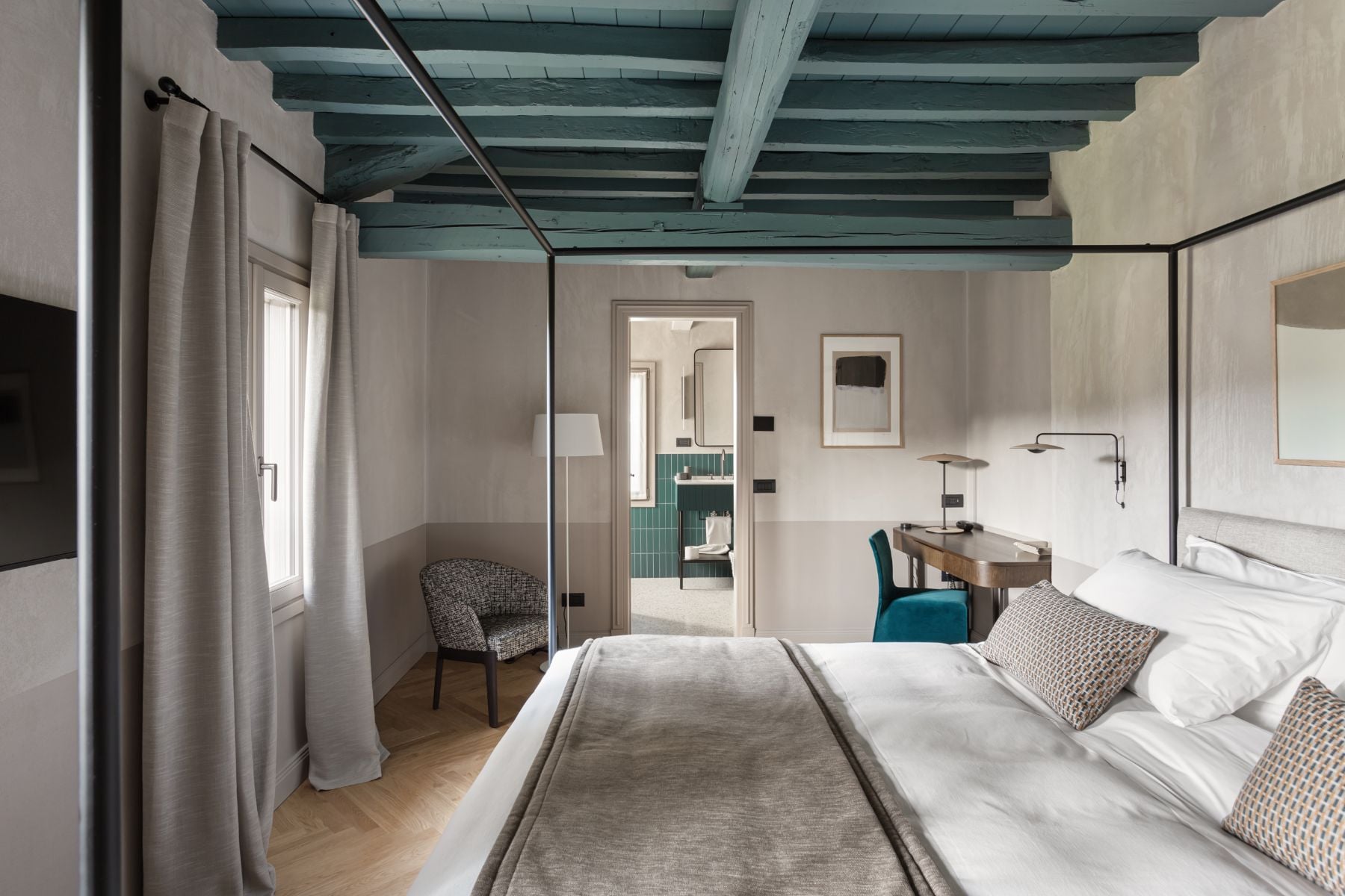 Roncolo 1888, Europe — Canopied bed with linen bedding in minimalist bedroom with exposed teal ceiling beams, ensuite bathroom, and natural light from windows.