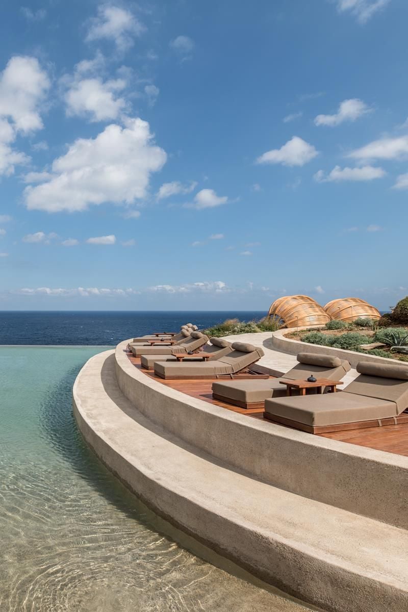 ACRO Suites, Europe — Curved concrete terrace with wooden loungers and golden dome structures overlooking turquoise infinity pool and open ocean.