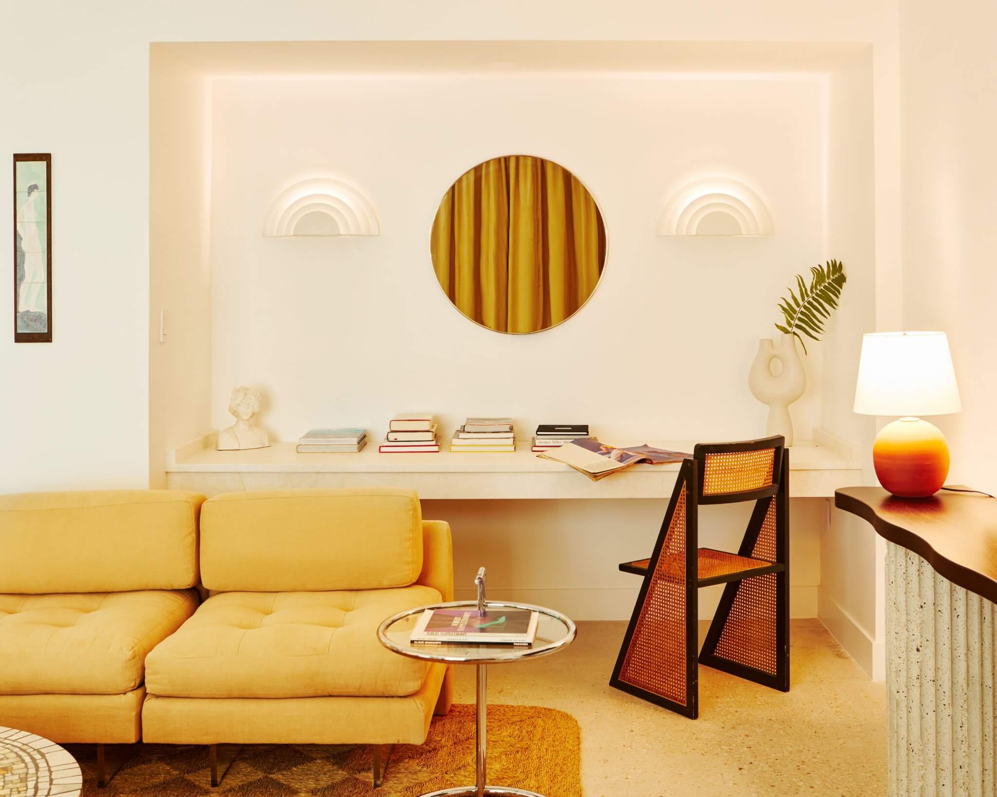 Palm Heights, North America — Minimalist living room with golden-yellow sectional sofa, curved rattan chair, cream built-in shelving, circular brass mirror, and warm ambient lighting.