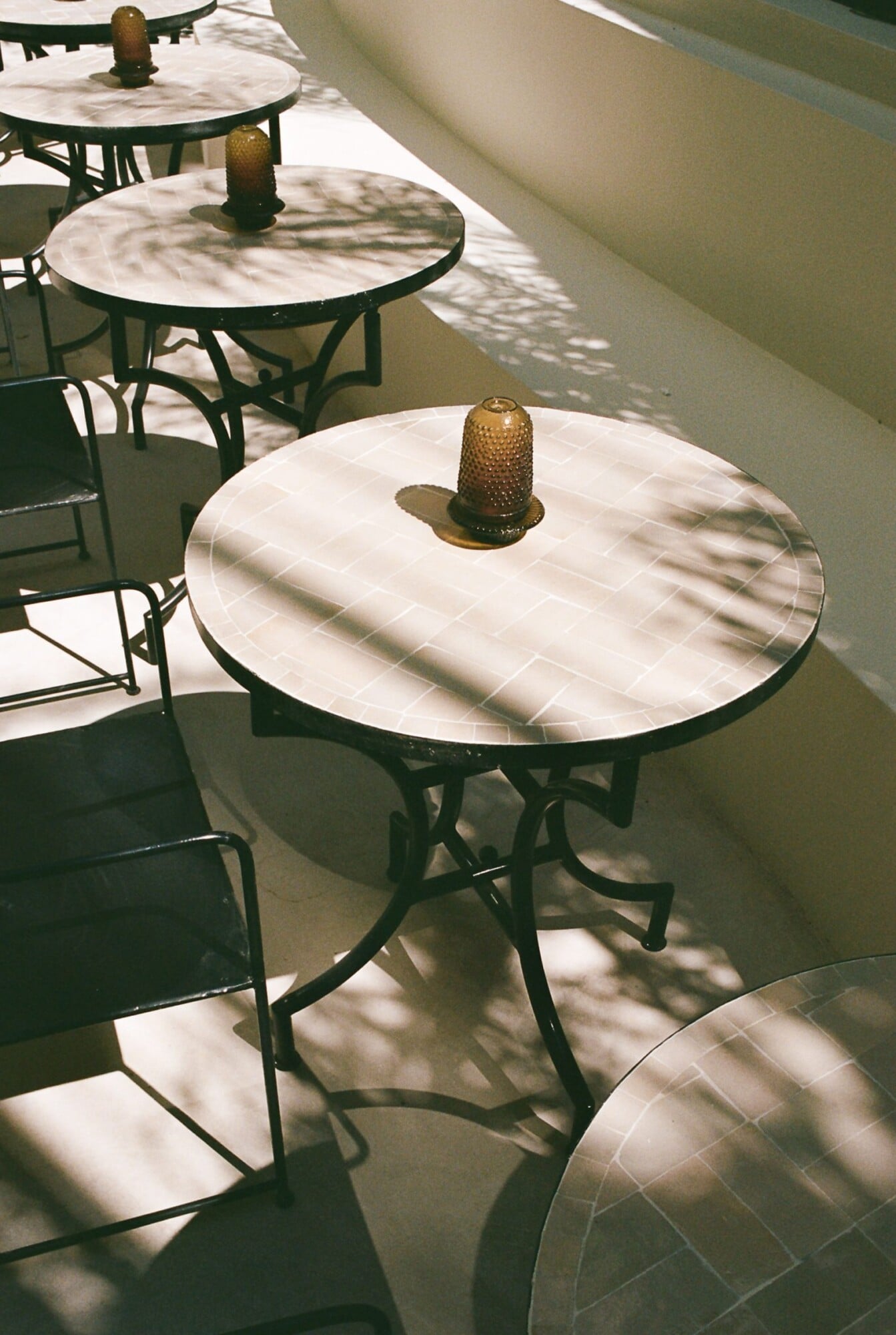 Apollo Palm, Europe — Marble-top bistro tables with brass bell-shaped centerpieces casting geometric shadows in dappled sunlight through minimalist colonnade.