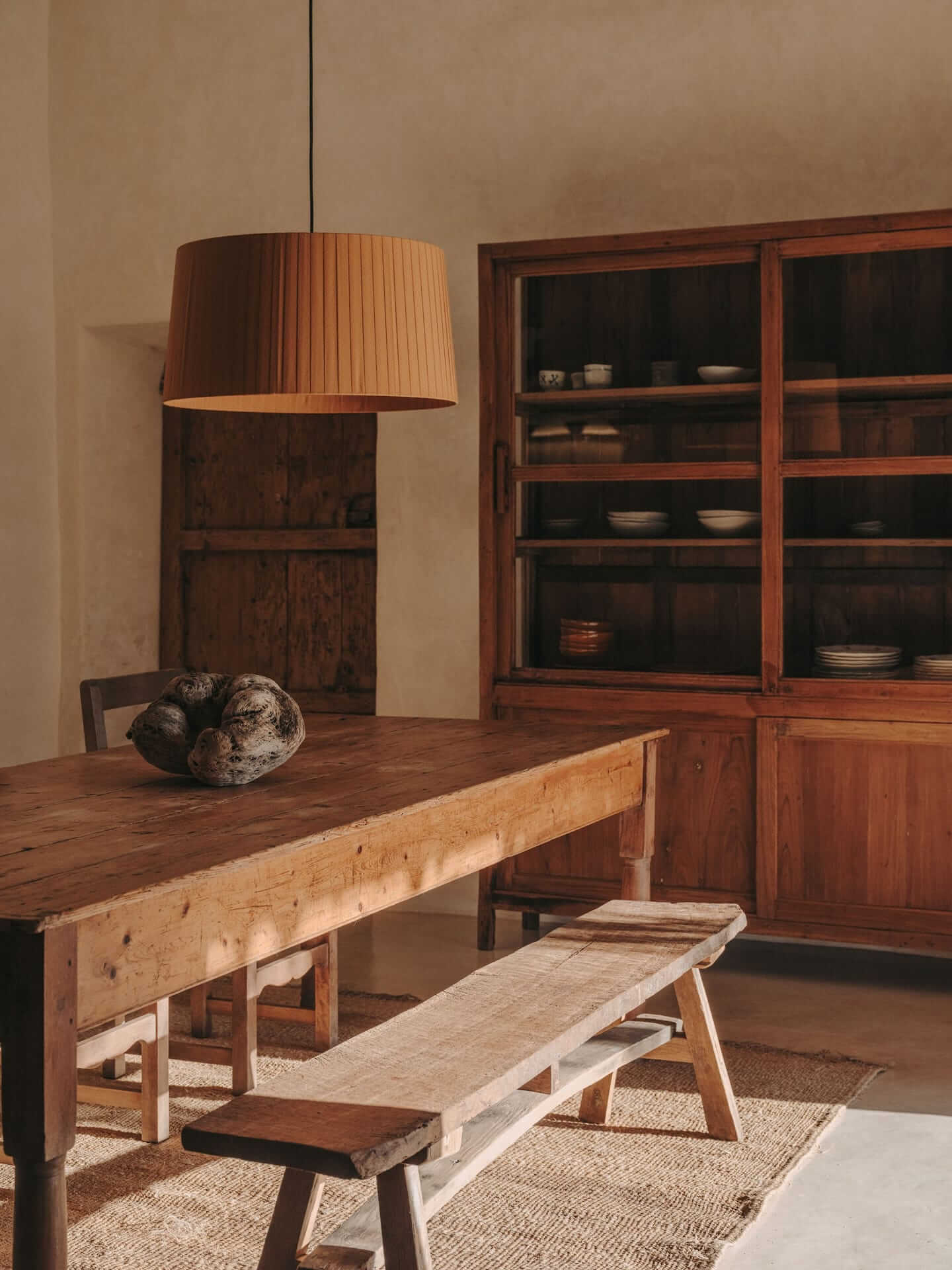 Can Lluïssó, Balearic Islands, Spain — Warm wooden dining table with natural light, terracotta pendant lamp, and glass-fronted cabinet in minimalist Mediterranean interior.