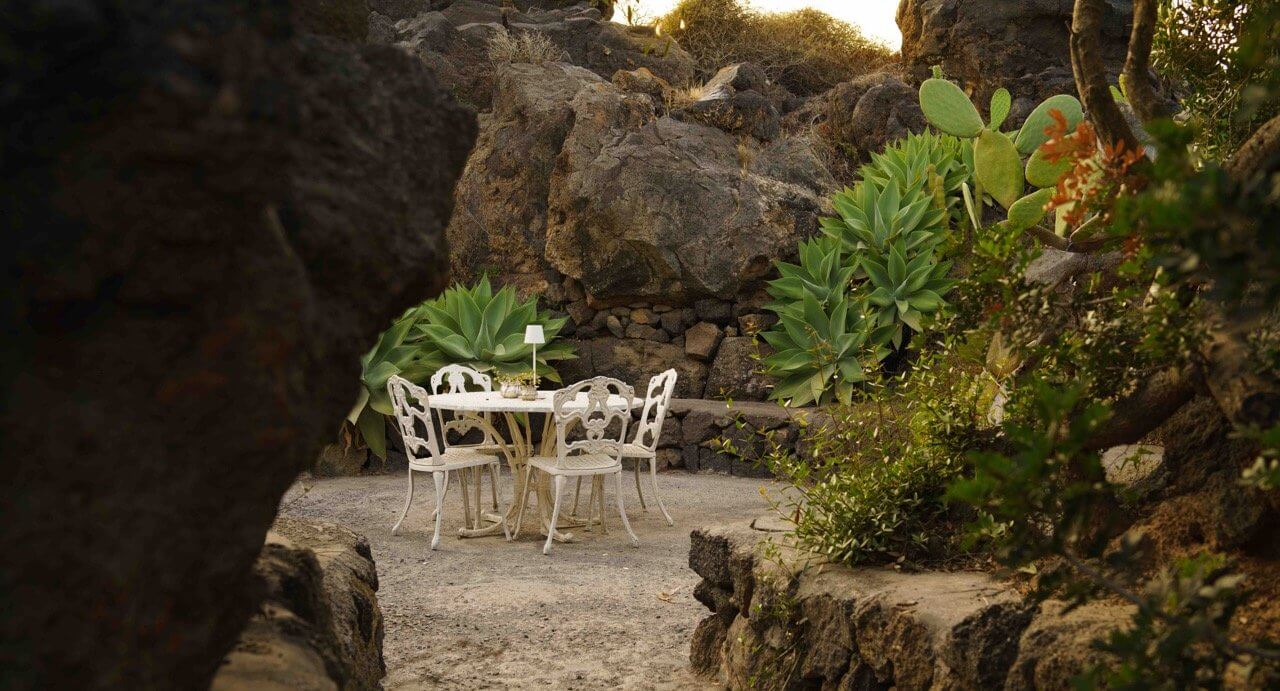 Parco Dei Sesi, Europe — White metal chairs and small table nestled in rocky stone alcove surrounded by succulents and coastal vegetation at golden hour.