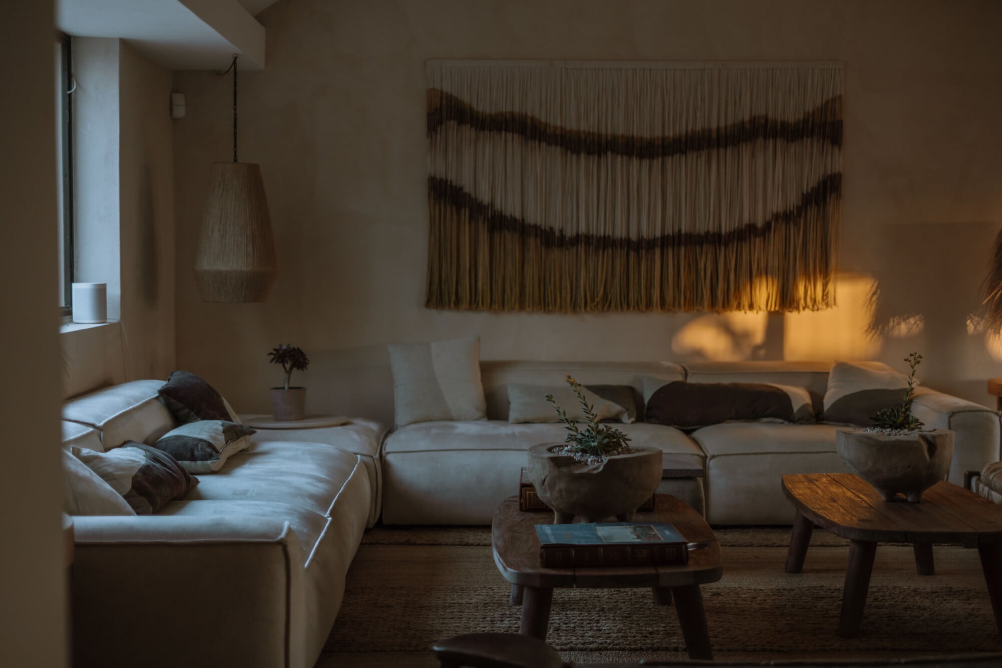 Tenuta Negroamaro, Europe — Minimalist living room with cream modular seating, woven wall tapestry, wooden side tables, potted plants, and warm afternoon light casting shadows across neutral tones.