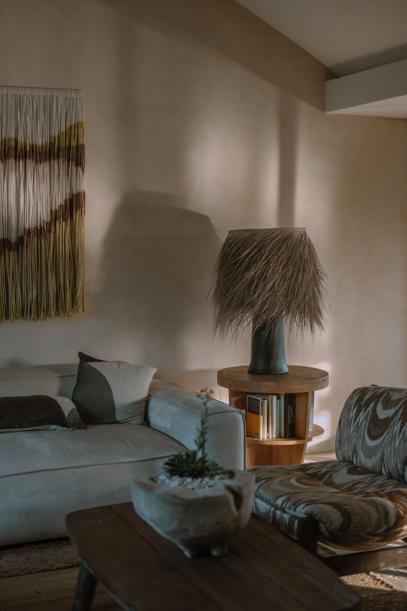 Tenuta Negroamaro, Europe — Minimalist living room with natural light, cream linen sofa, wooden side table with fringe lamp, woven wall hanging, and sculptural furnishings in warm earth tones.