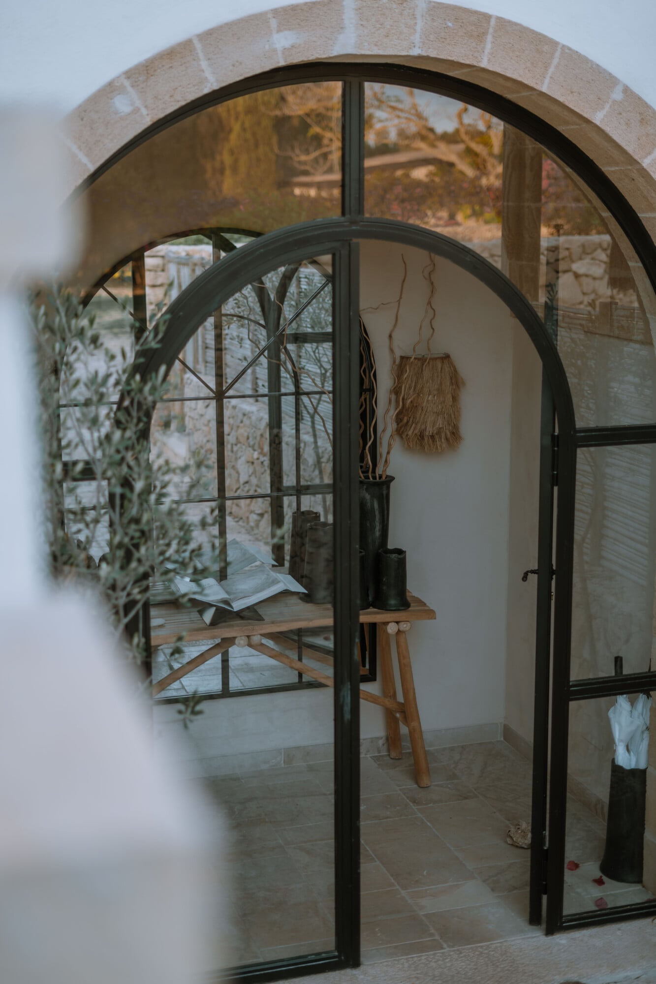 Tenuta Negroamaro, Europe — Arched doorway with black steel frame opening to minimalist interior, terracotta floors, dried botanicals, natural light through gridded windows.