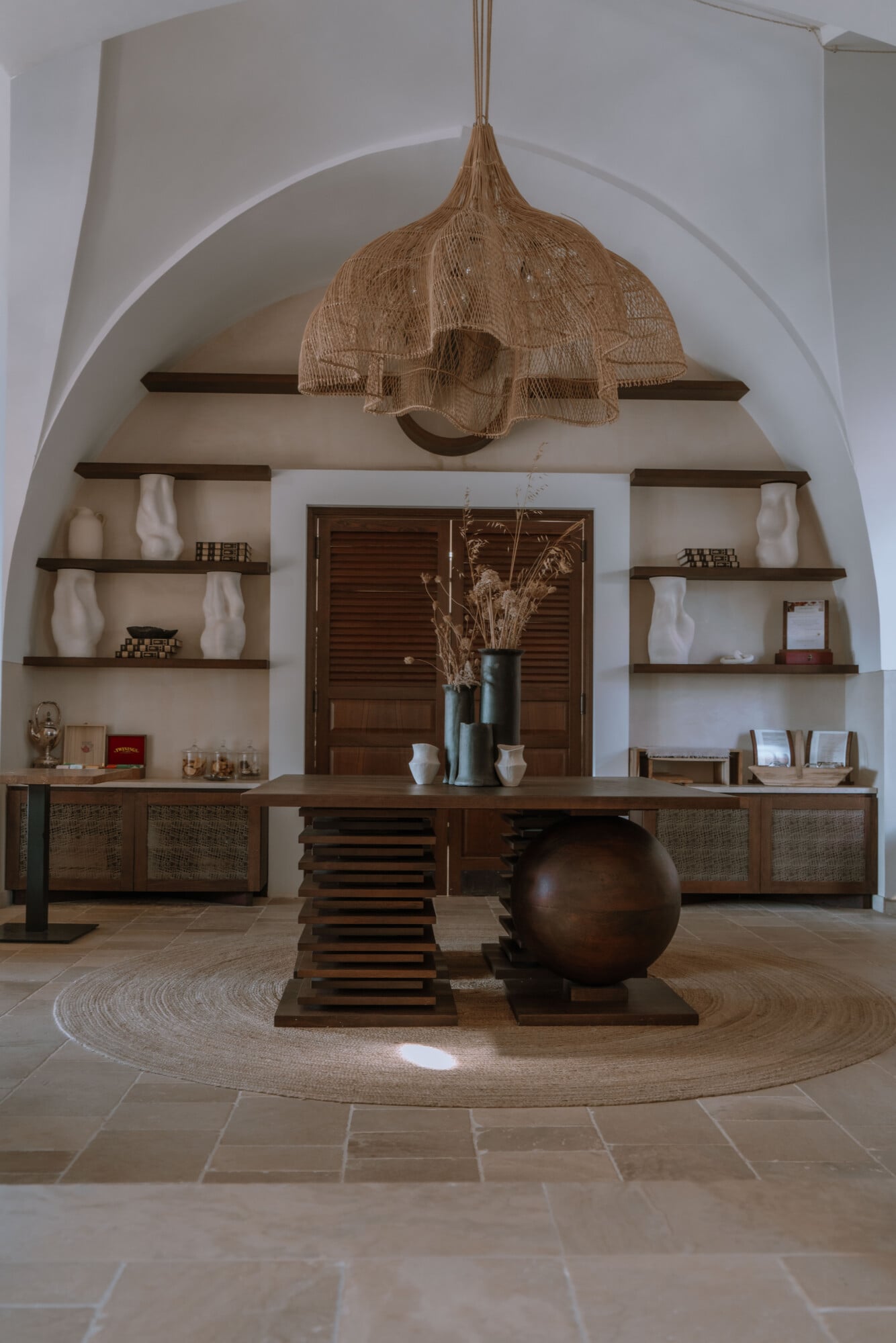 Tenuta Negroamaro, Europe — Arched alcove with woven pendant light, wooden shelving, bronze sculptural sphere, and linen round rug in minimalist hospitality interior.