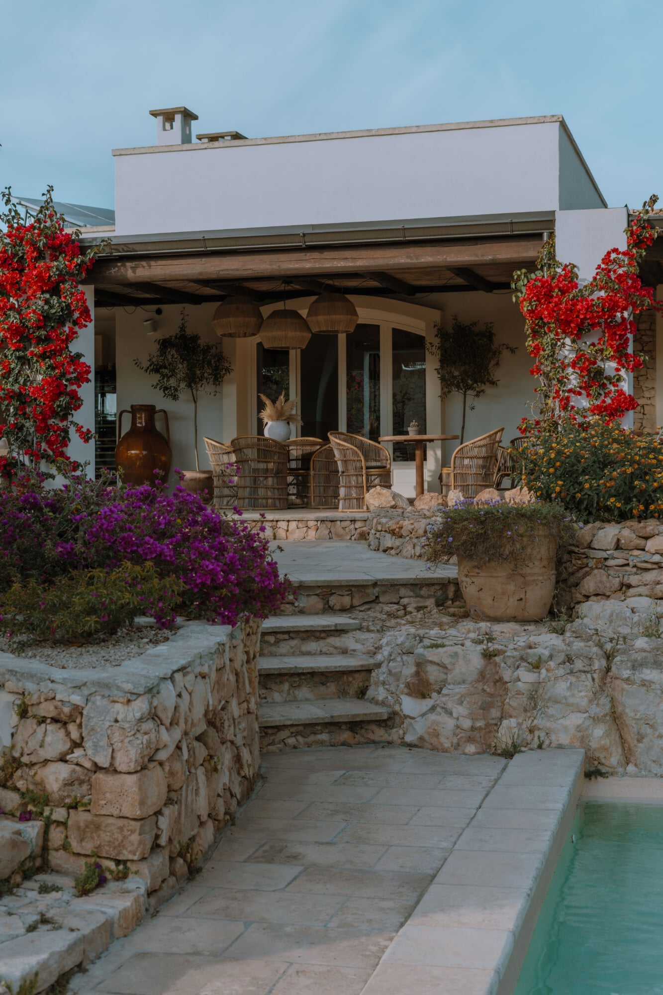 Tenuta Negroamaro, Europe — Modern white villa with wooden pergola, terracotta floors, vibrant bougainvillea and climbing flowers, stone retaining walls, woven chairs, pool edge, Mediterranean architecture.