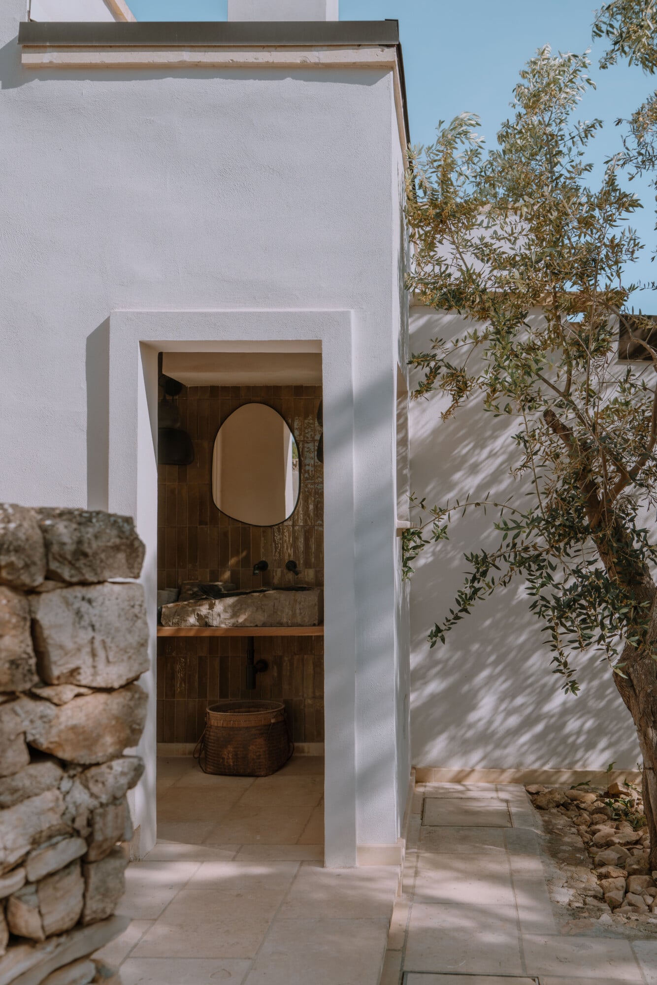 Tenuta Negroamaro, Europe — Whitewashed bathroom entrance with arched mirror, dark wood interior, natural light casting shadows from olive tree, minimalist Mediterranean design.