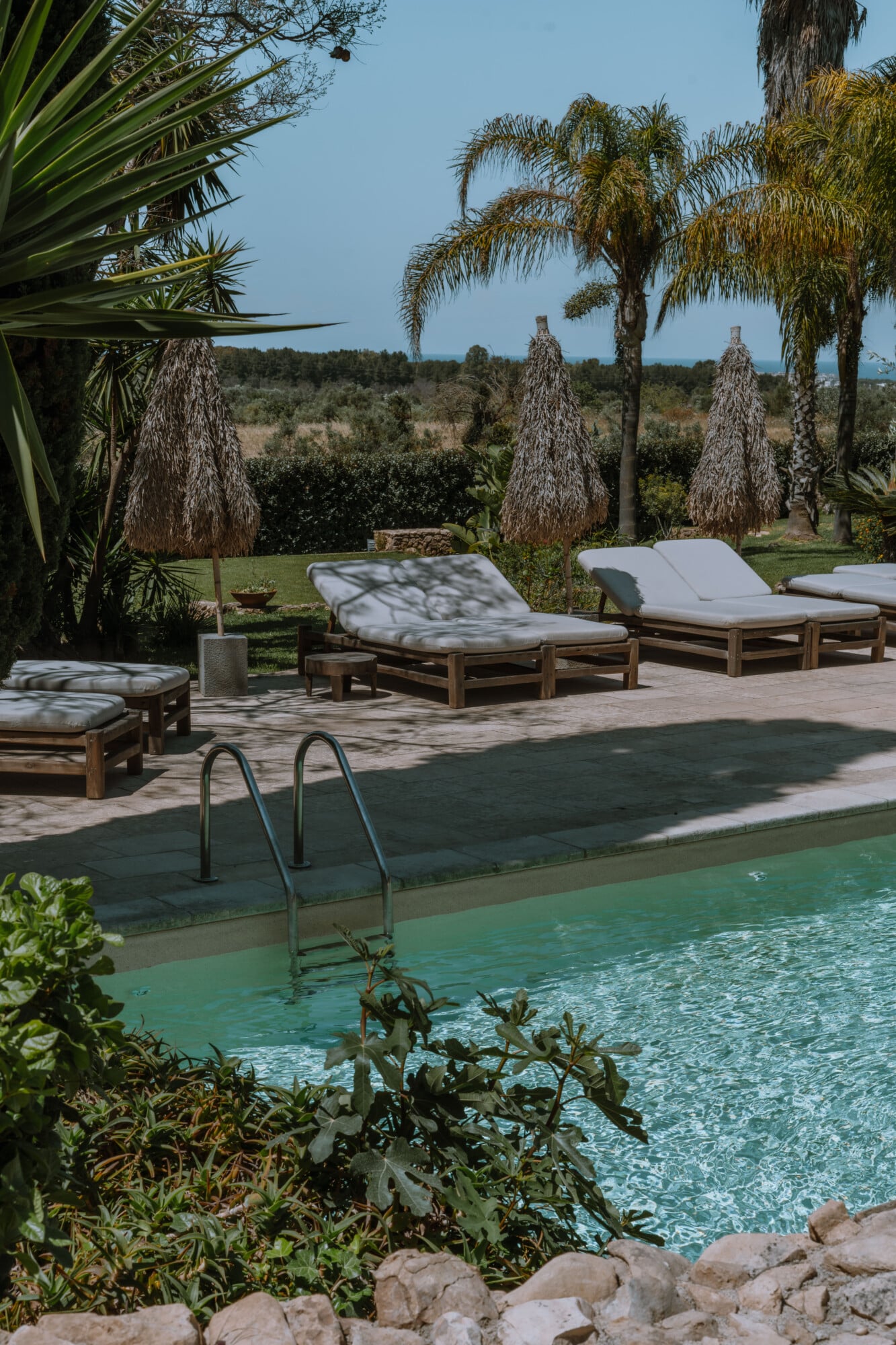 Tenuta Negroamaro, Europe — Poolside lounge with teak daybeds, white cushions, palm trees, and manicured gardens overlooking countryside under clear sky.