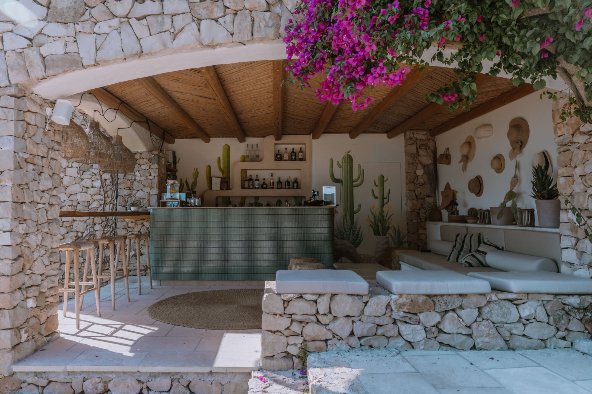 The Cactus Bar — Cactus Bar, Europe — Stone terrace with wooden beam ceiling, green tiled bar counter, saguaro cacti, magenta bougainvillea, and cushioned stone seating in Mediterranean design.