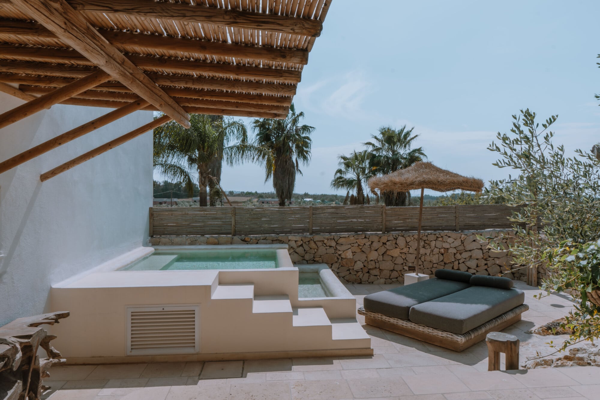 Tenuta Negroamaro, Europe — Minimalist poolside terrace with wooden pergola, stone walls, olive trees, and woven sun shade overlooking agricultural landscape.