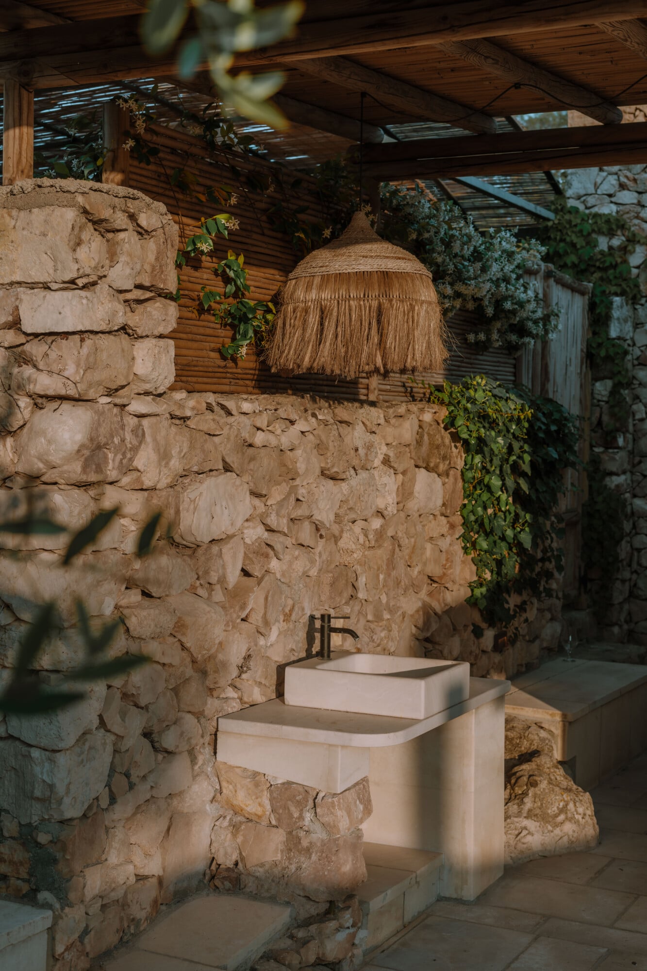 Tenuta Negroamaro, Europe — Stone courtyard with woven pendant lamp, modern white sink pedestal, wooden pergola, climbing ivy, warm natural light.