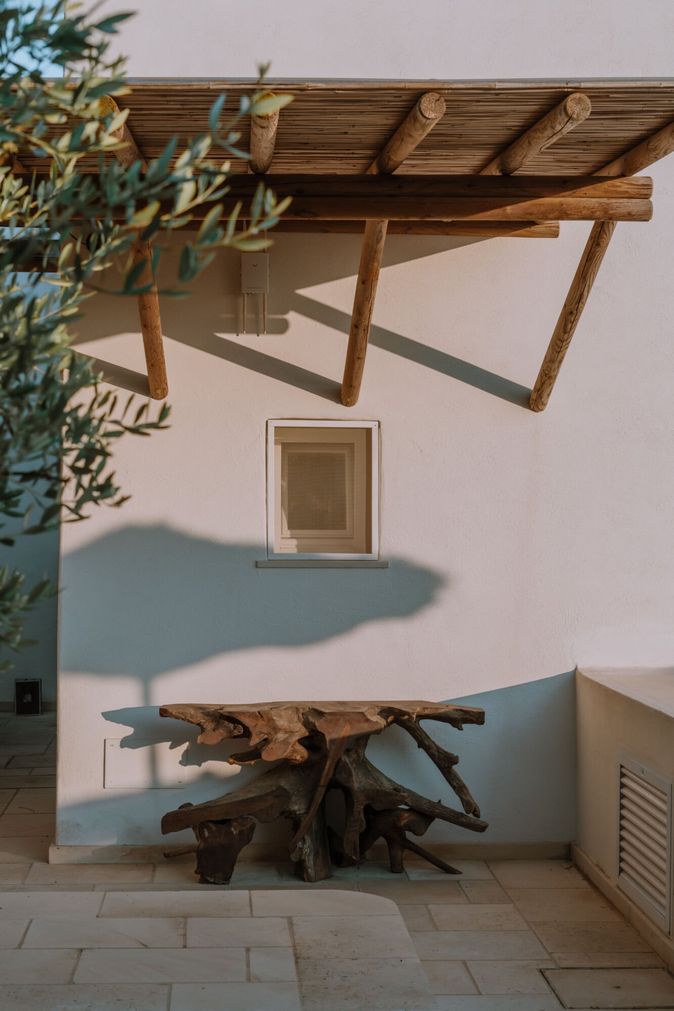 Tenuta Negroamaro, Europe — Pergola with weathered timber and driftwood table casting shadows on whitewashed courtyard wall with window.