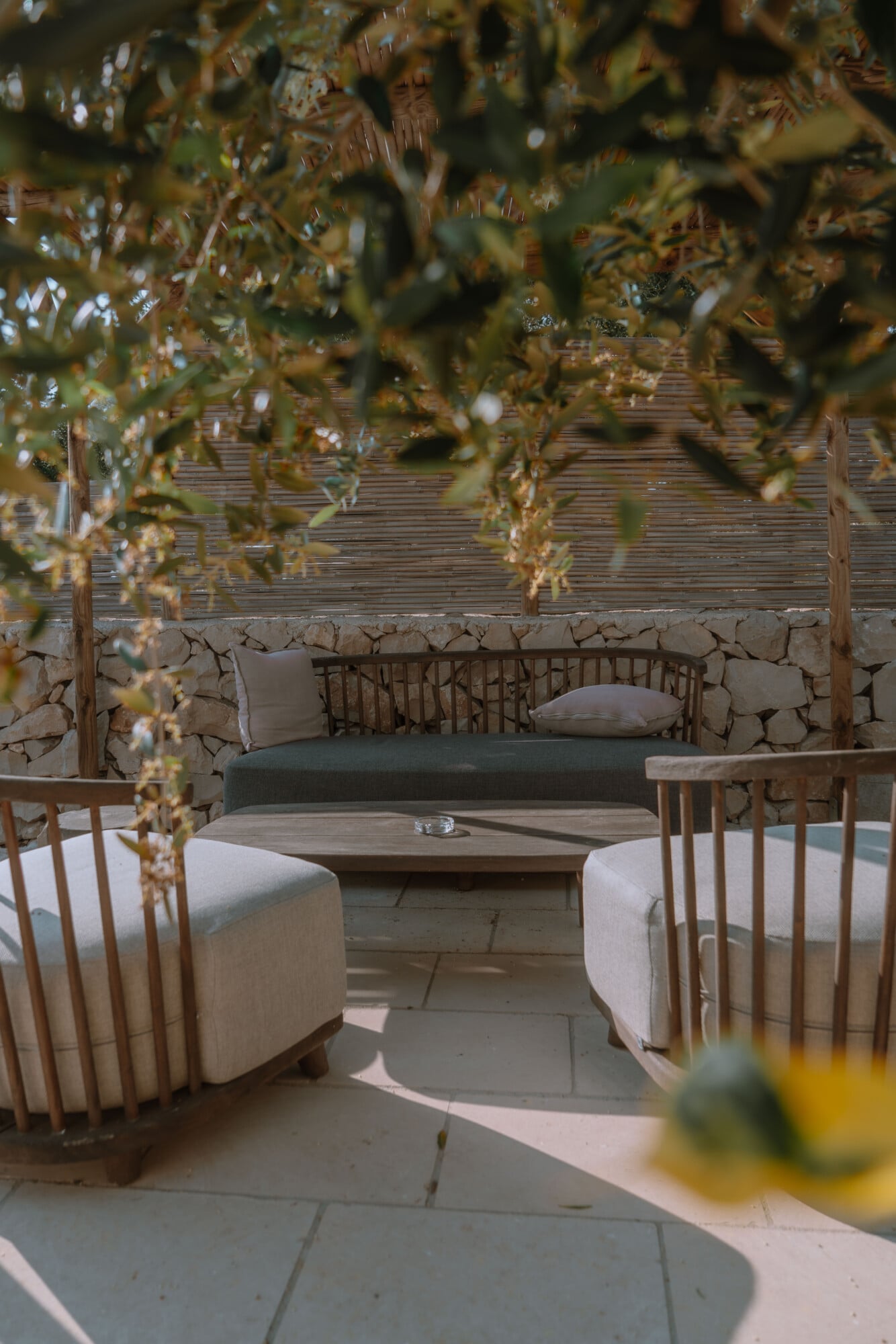 Tenuta Negroamaro, Europe — Courtyard seating area with curved linen ottomans, wooden chairs, stone walls, and dried climbing vines in natural light.