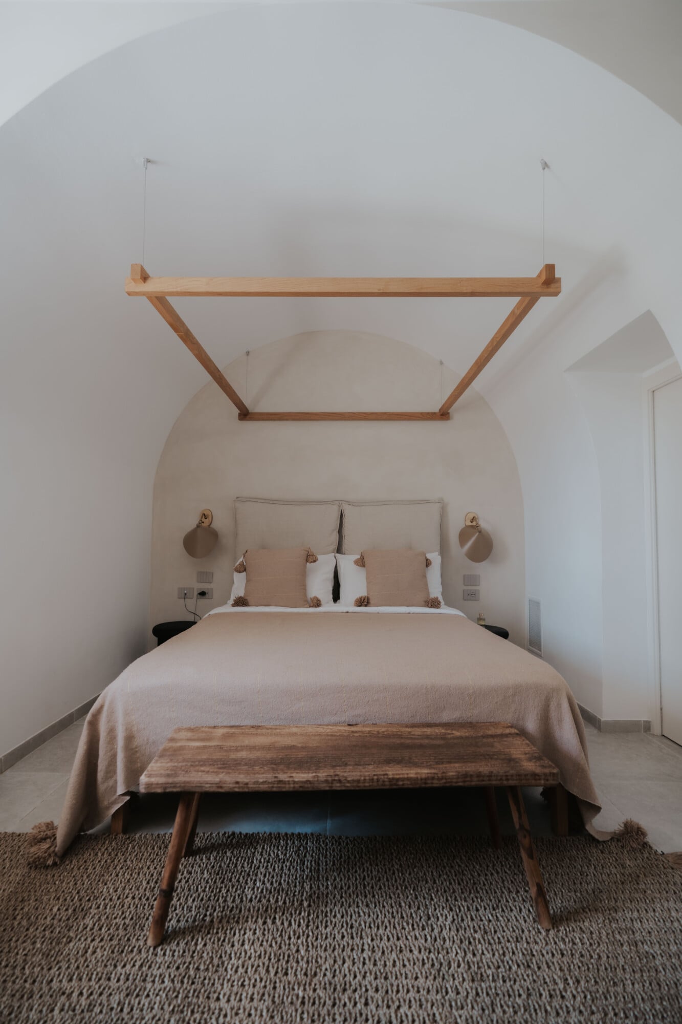 Tenuta Negroamaro, Europe — Minimalist bedroom with arched alcove, wooden canopy frame, neutral linens, terracotta sconces, and natural light in modern Mediterranean design.