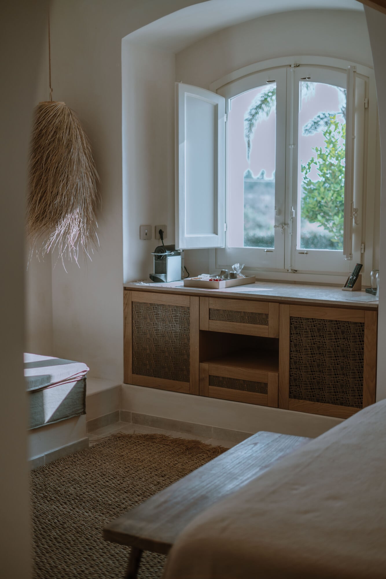 Tenuta Negroamaro, Europe — Arched stone alcove with wooden cabinetry, marble countertop, tall window overlooking gardens, woven pendant light, natural materials and warm minimalist interiors.