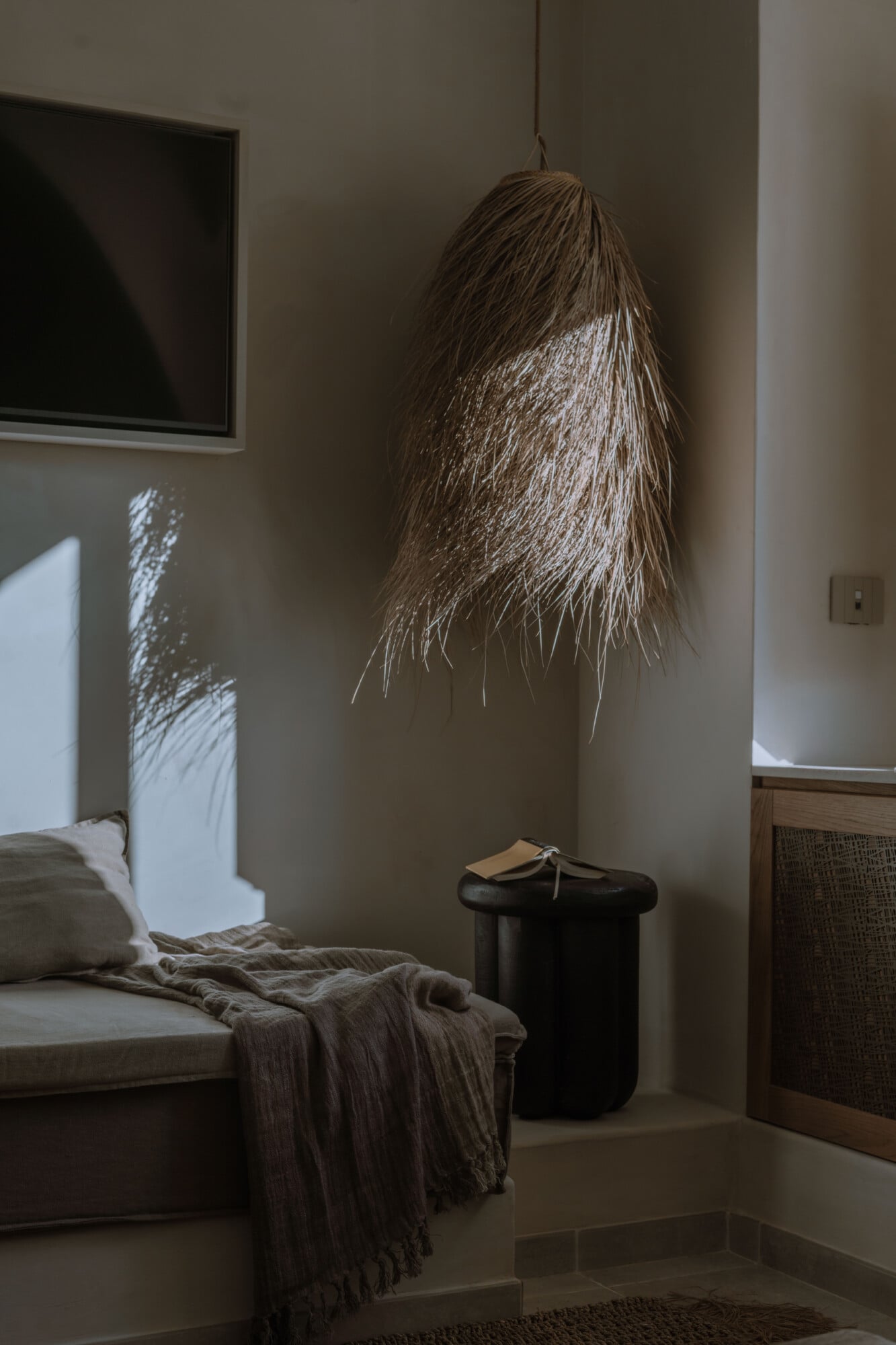 Tenuta Negroamaro, Europe — Minimalist bedroom with woven pendant lamp, natural light casting shadows on neutral walls, earthy textiles, and black cylindrical side table.