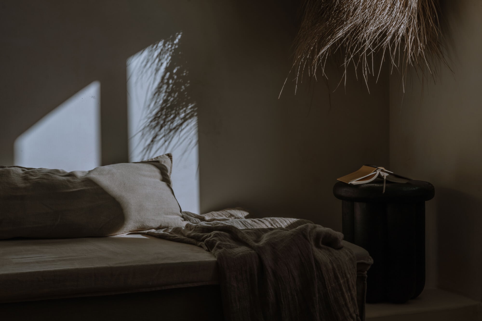 Tenuta Negroamaro, Europe — Minimalist bedroom with natural light casting shadows across white pillow and linen bedding, dark wooden nightstand, neutral walls.