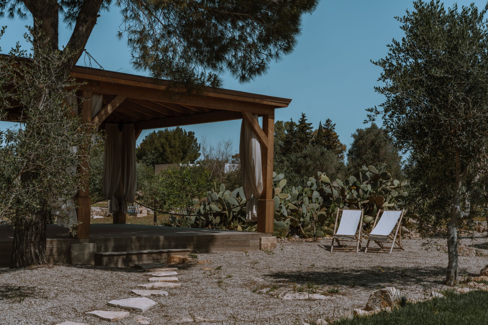 Yoga Europe — wooden pergola with draped curtains casting shadows on gravel, flanked by olive trees and deck chairs, Mediterranean landscape beyond.