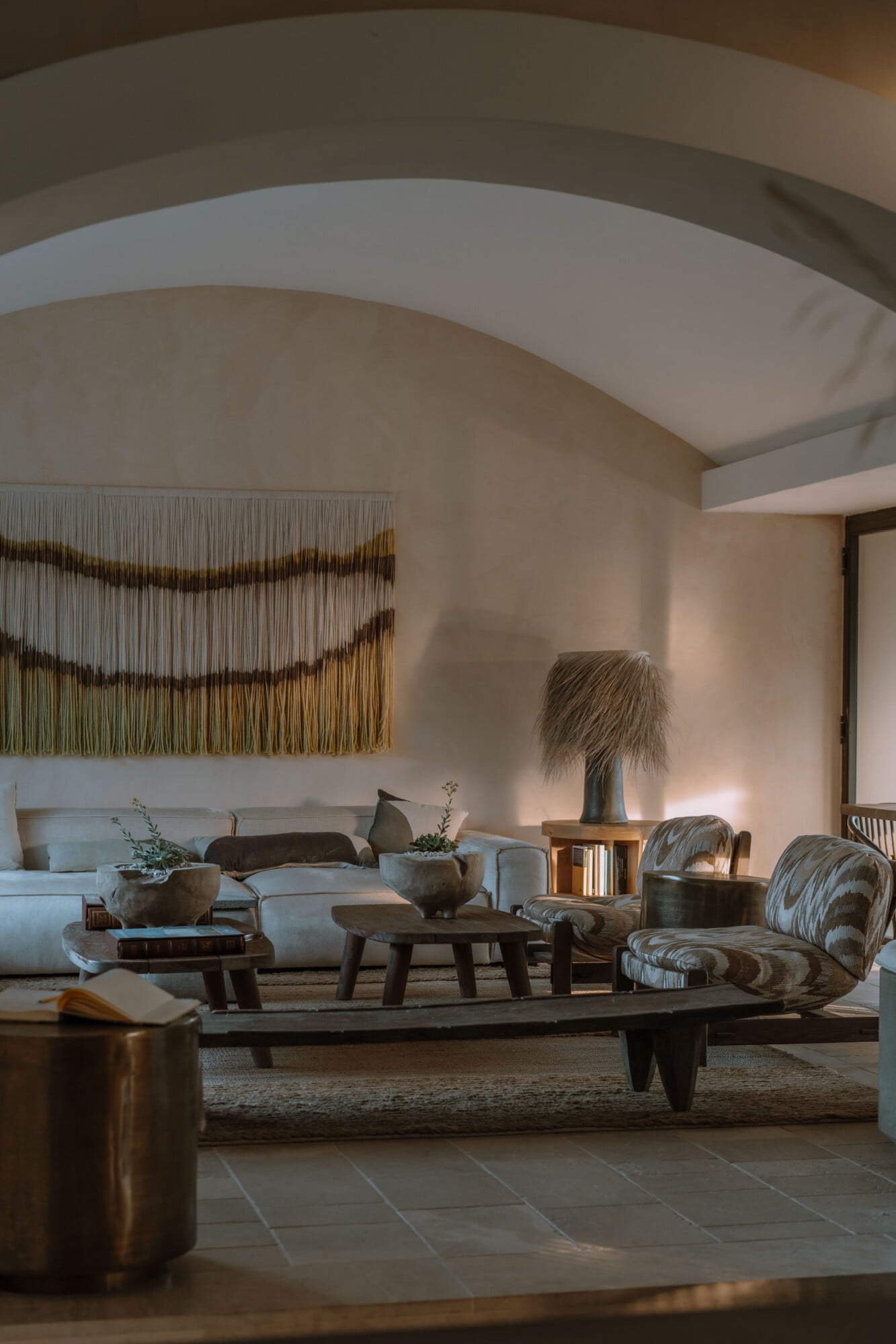 Tenuta Negroamaro, Europe — Curved white ceiling arches over minimalist living room with cream sofa, patterned chairs, wooden tables, macramé wall hanging, and warm natural light.
