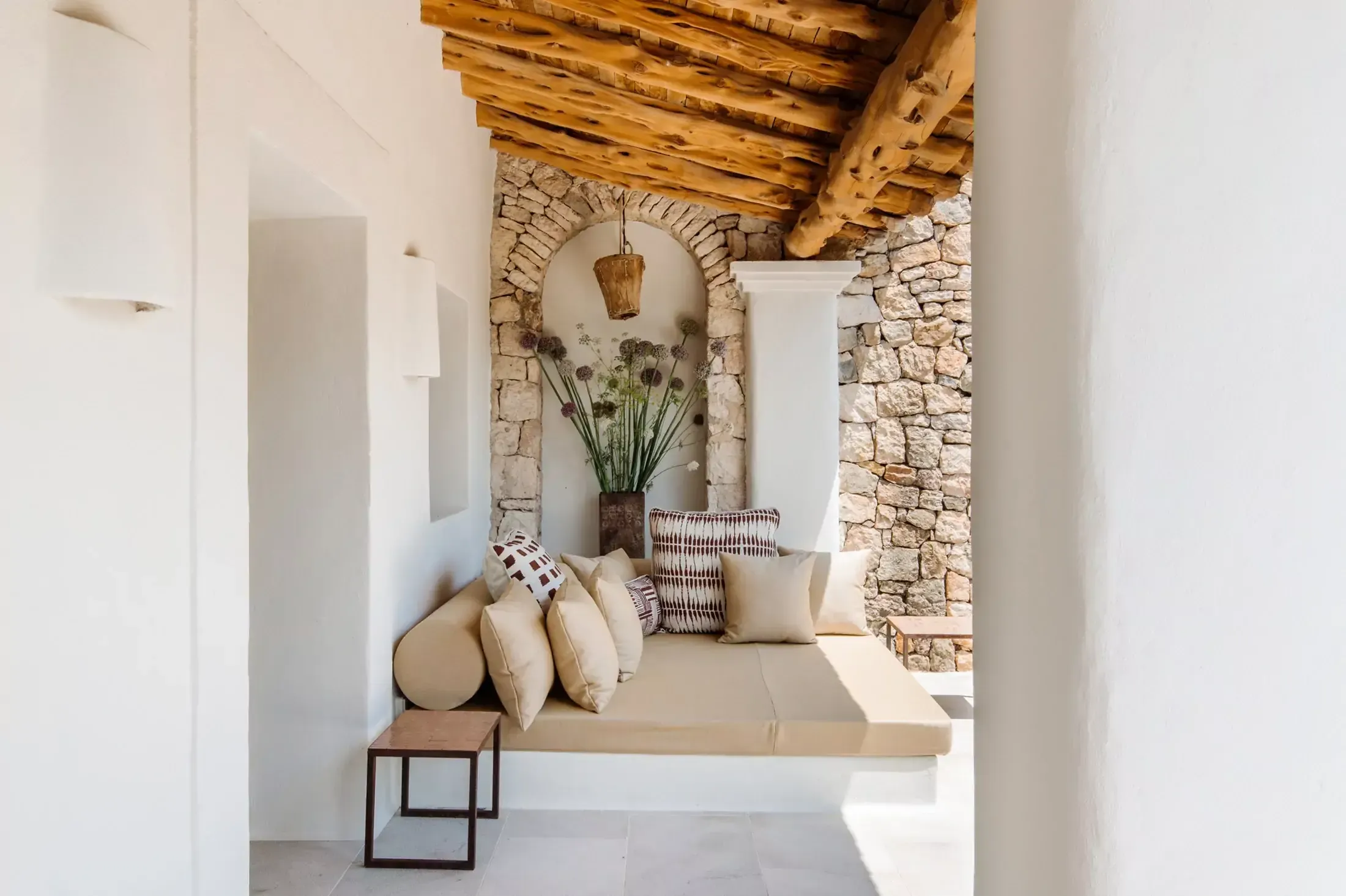 Finca La Inika, Balearic Islands, Spain — Stone archway with beige daybed, wooden ceiling beams, decorative pillows, potted grasses, and woven pendant light in minimalist Mediterranean interior.