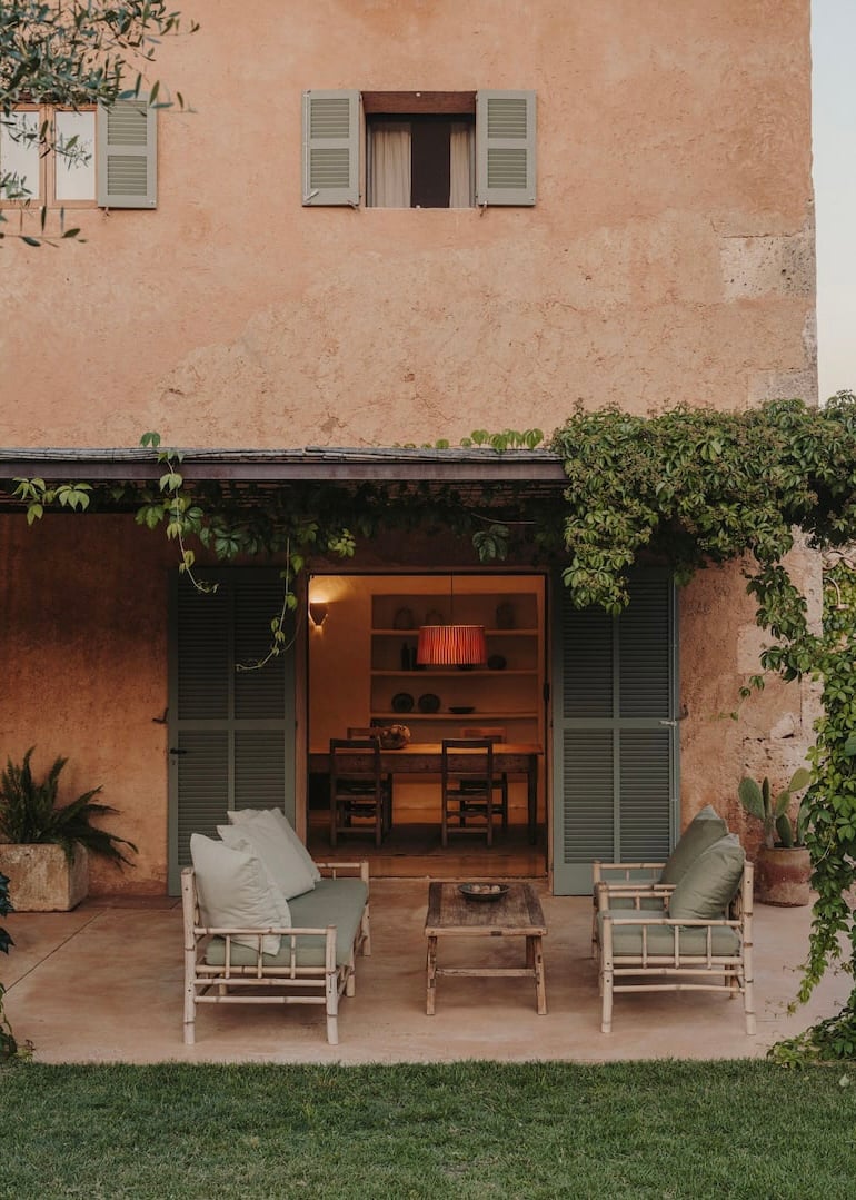 Can Lluïssó, Balearic Islands, Spain — Terracotta courtyard with whitewashed seating, dark timber pergola, and warm interior light beyond open doors.