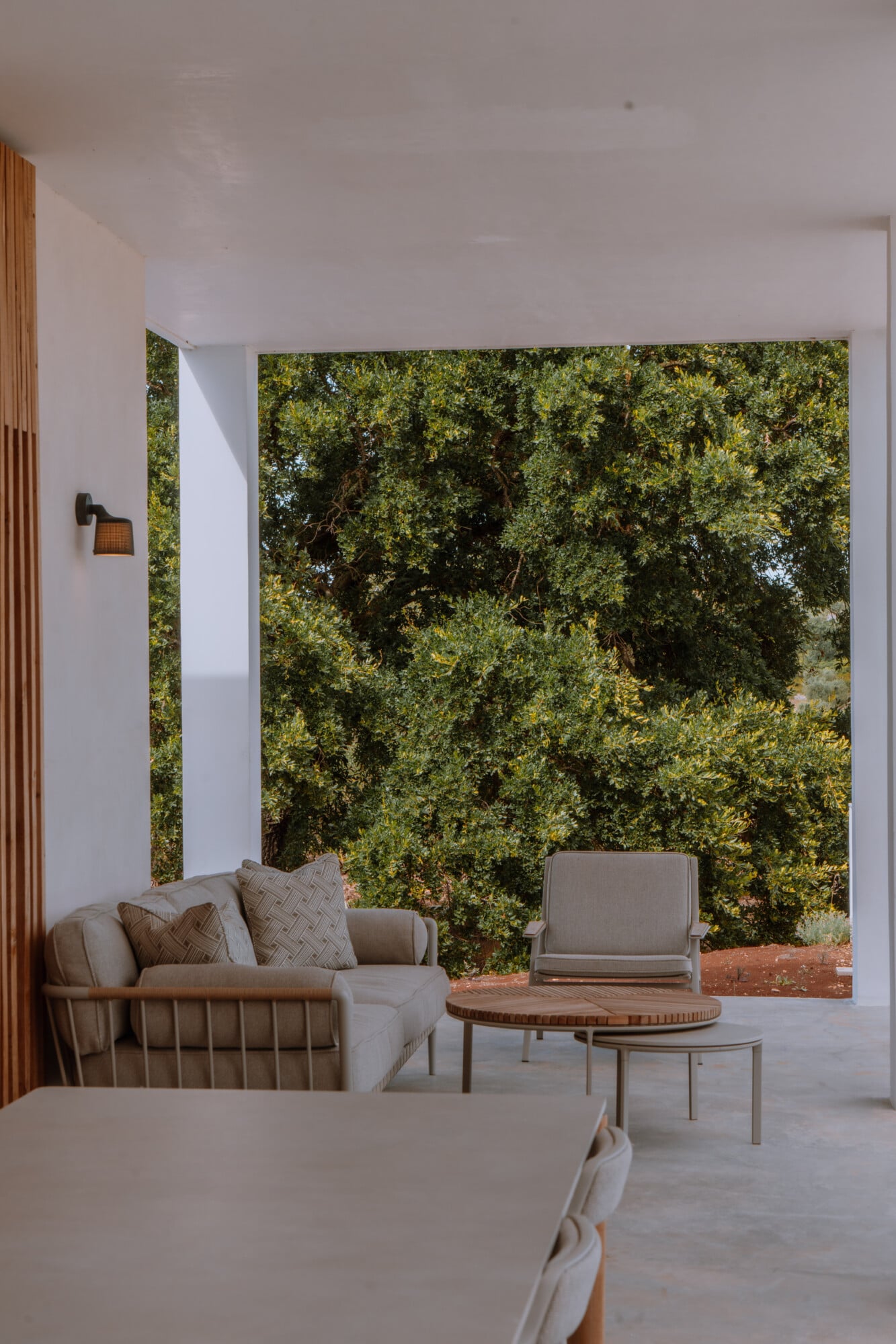 Villa Quercia, Italy — Minimalist lounge with beige linen seating, wooden side tables, and floor-to-ceiling view of dense green foliage and terracotta courtyard.