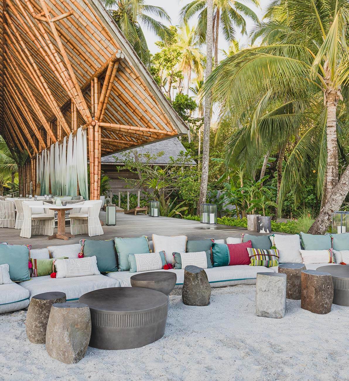 The Terrazza — Beachfront lounge with curved modular seating, stone fire pit, palm trees, and teak pavilion architecture in soft coastal light.