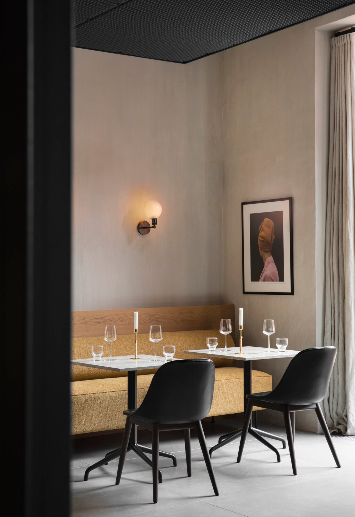 The Audo, Europe — Minimalist dining alcove with gold-upholstered banquette, black chairs, white marble tables, textured black ceiling, warm sconce lighting, and framed portrait artwork.