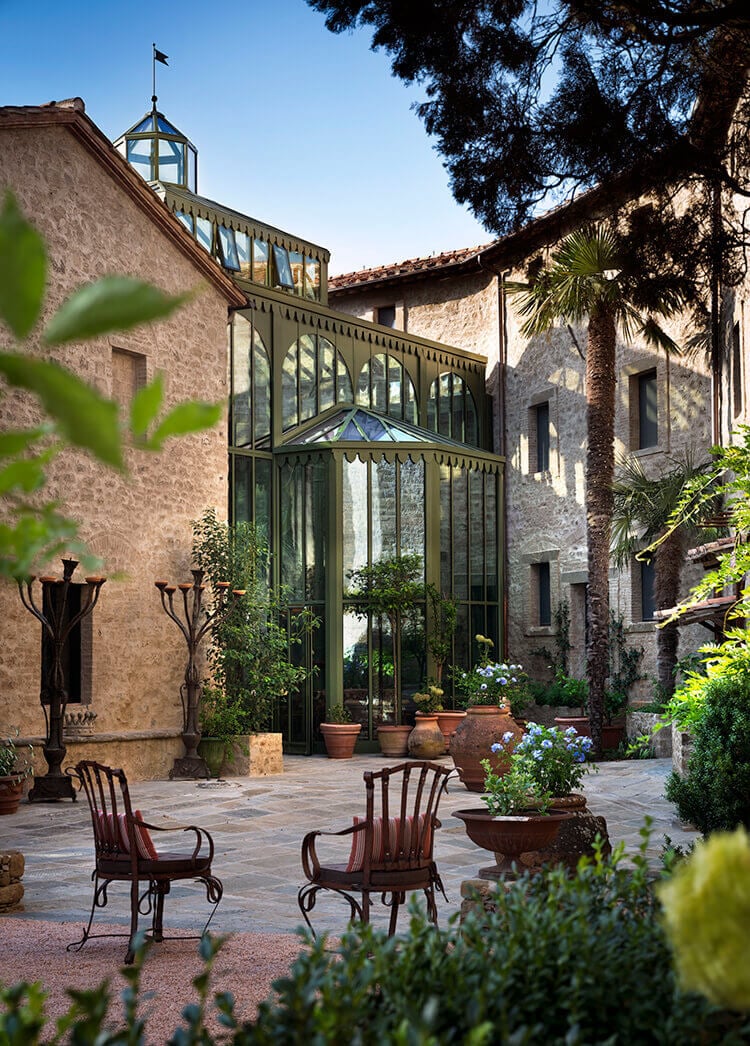 Castello di Reschio, Italy — Stone courtyard with green-framed conservatory, terracotta planters, wooden chairs, palm trees, and ochre-hued Renaissance architecture under clear sky.