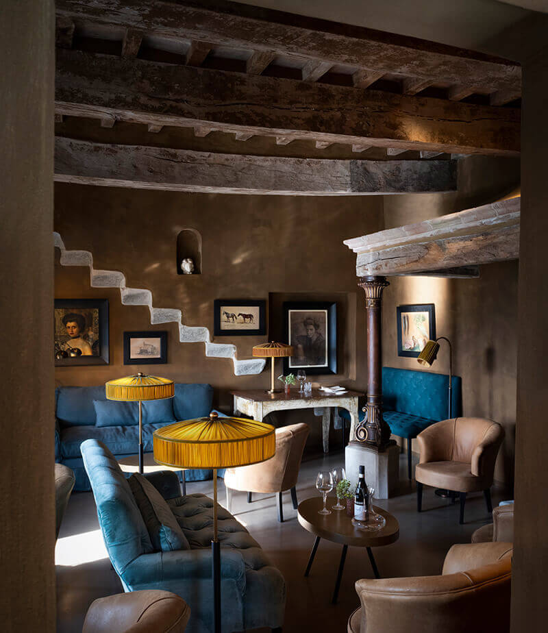 Castello di Reschio, Europe — Rustic stone ceiling beams frame a luxe lounge with blue velvet seating, golden side tables, terracotta walls, and curated artwork.