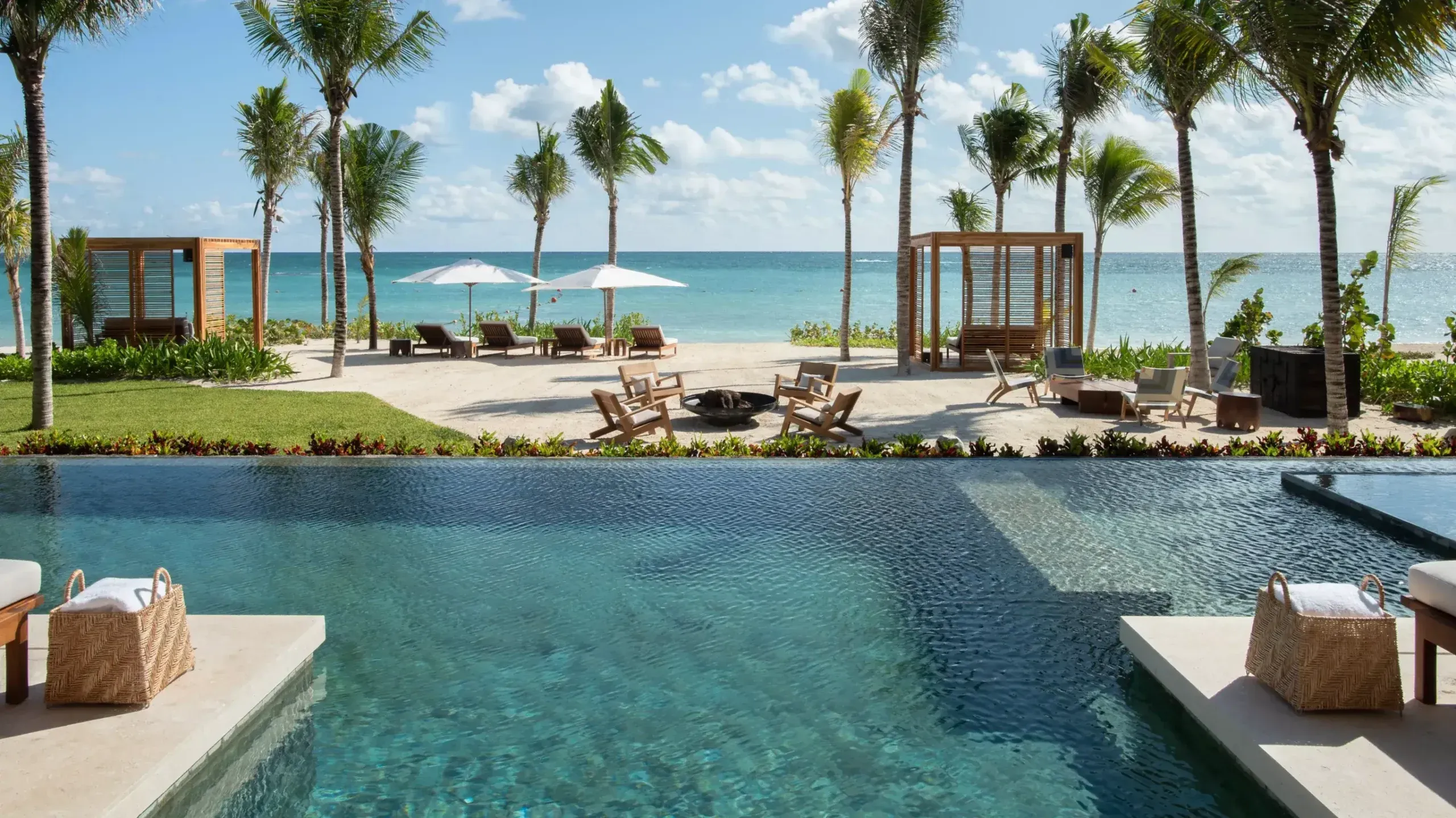 Rosewood Mayakoba, Mexico — Infinity pool overlooking Caribbean waters with palm-lined beach, wooden loungers, fire pit, and modernist pavilions at sunset.
