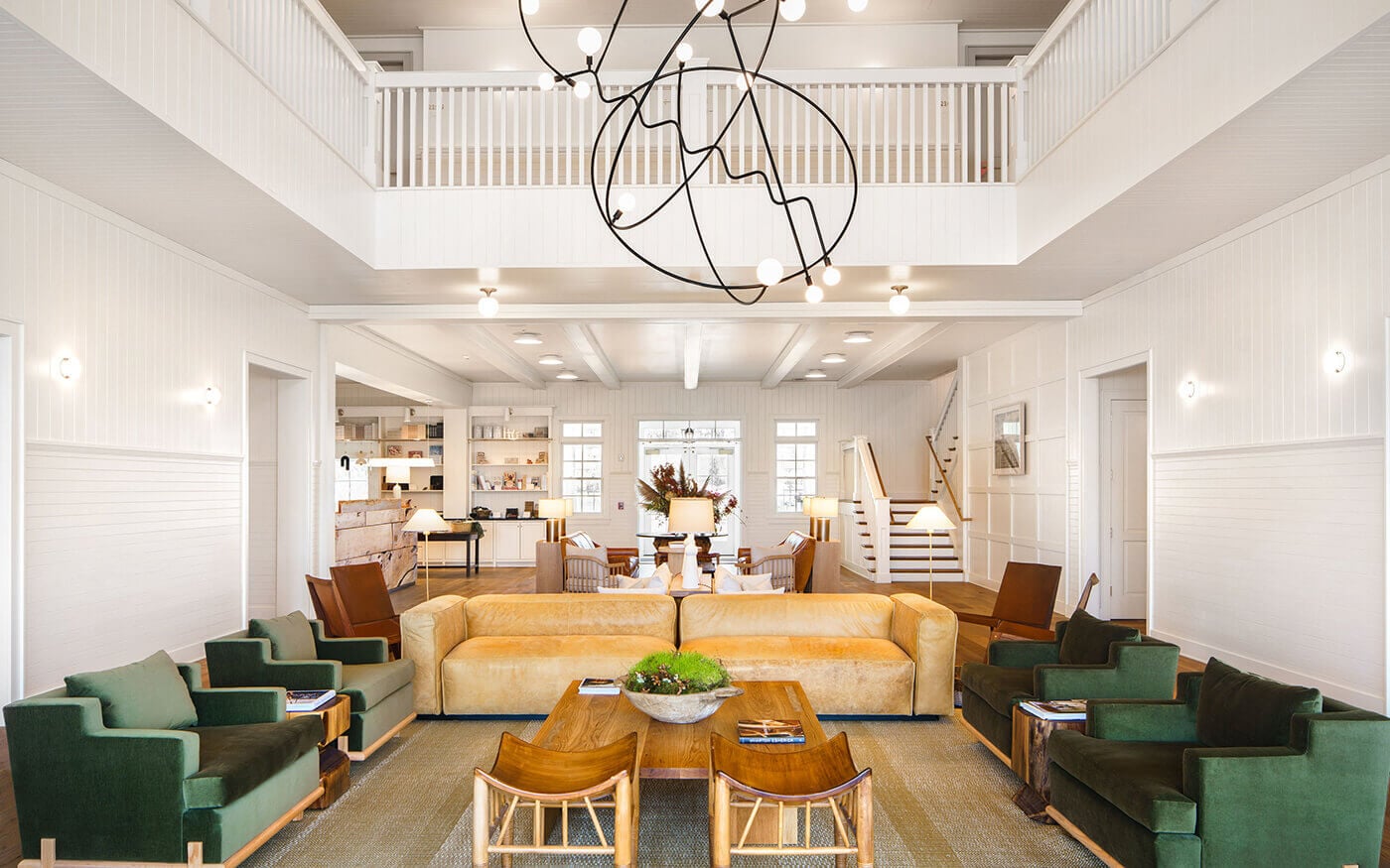 The Lake House — Lake House, North America — Luminous double-height lobby with cream shiplap walls, golden leather sofa, green velvet seating, oak coffee table, abstract black chandelier, and skylit clerestory windows.