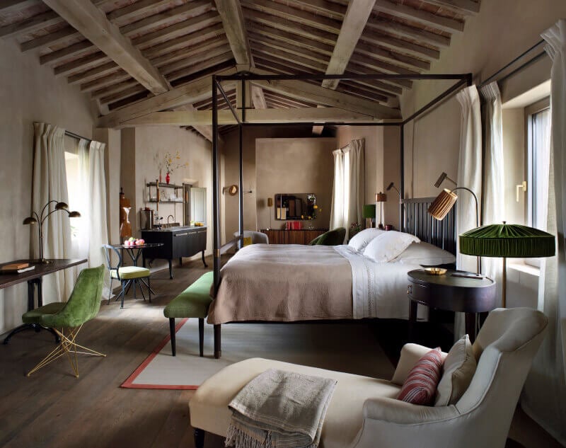 Castello di Reschio, Umbria, Italy — Canopy bed in stone-walled bedroom with exposed timber ceiling, green velvet seating, and natural light from tall windows.