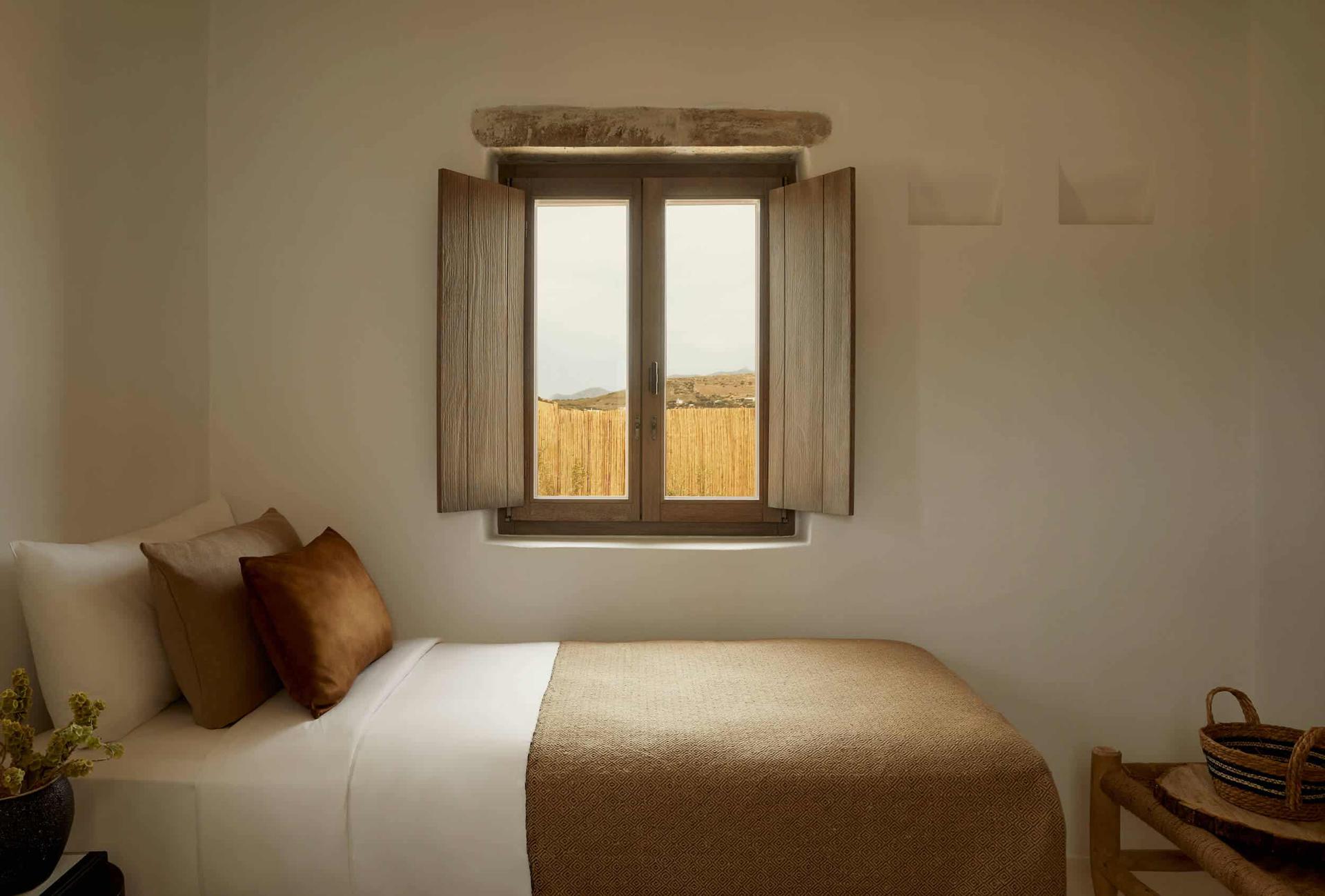 The Rooster, Europe — Minimalist bedroom with wooden shuttered window overlooking golden wheat fields, linen bedding, bronze silk pillows, terracotta pot, woven basket.