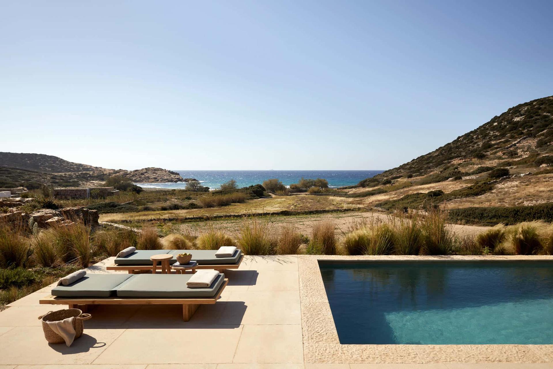 The Rooster, Europe — Limestone terrace with teak loungers, infinity pool overlooking Mediterranean cove, dry grasses and rocky hills, clear daylight.