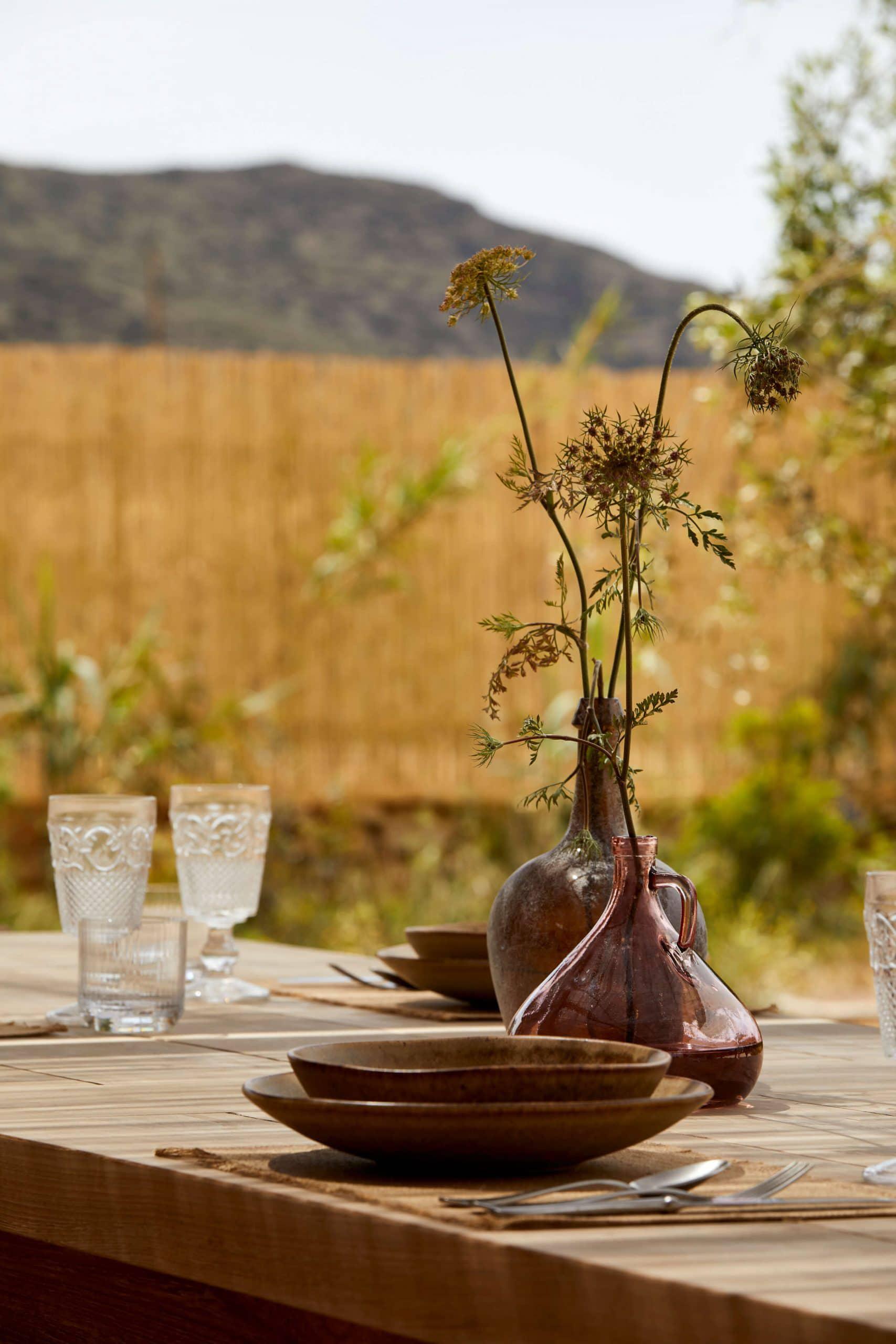 The Rooster, Europe — Outdoor dining table with dried flower arrangement in terracotta vessel, wooden bowls, glassware, mountain and golden field backdrop.