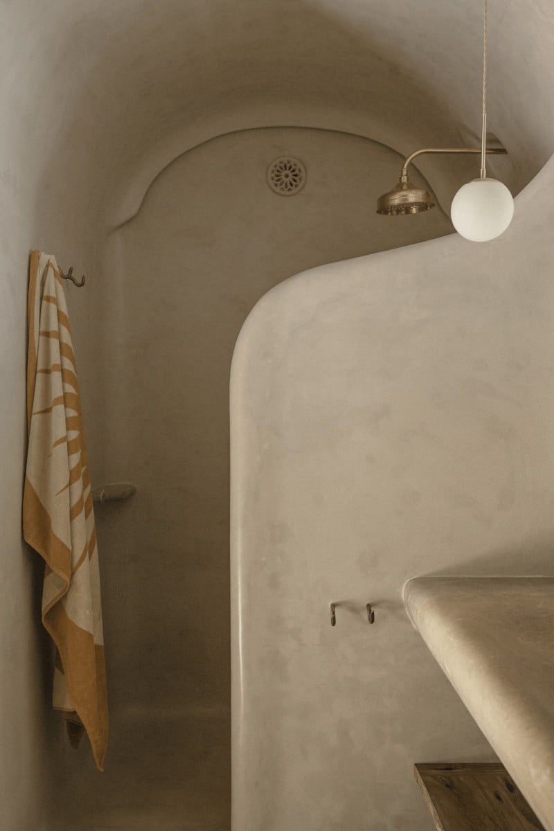 Hotel Corazón, Balearic Islands — Minimalist bathroom with curved plaster walls, brass shower fixture, white globe pendant, and ochre striped linen towel.