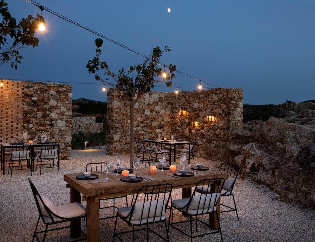 The Secret Garden  — Secret Garden, Europe — Stone pavilions with warm string lights and lanterns frame candlelit wooden dining table at dusk, moon rising above rocky Mediterranean landscape.