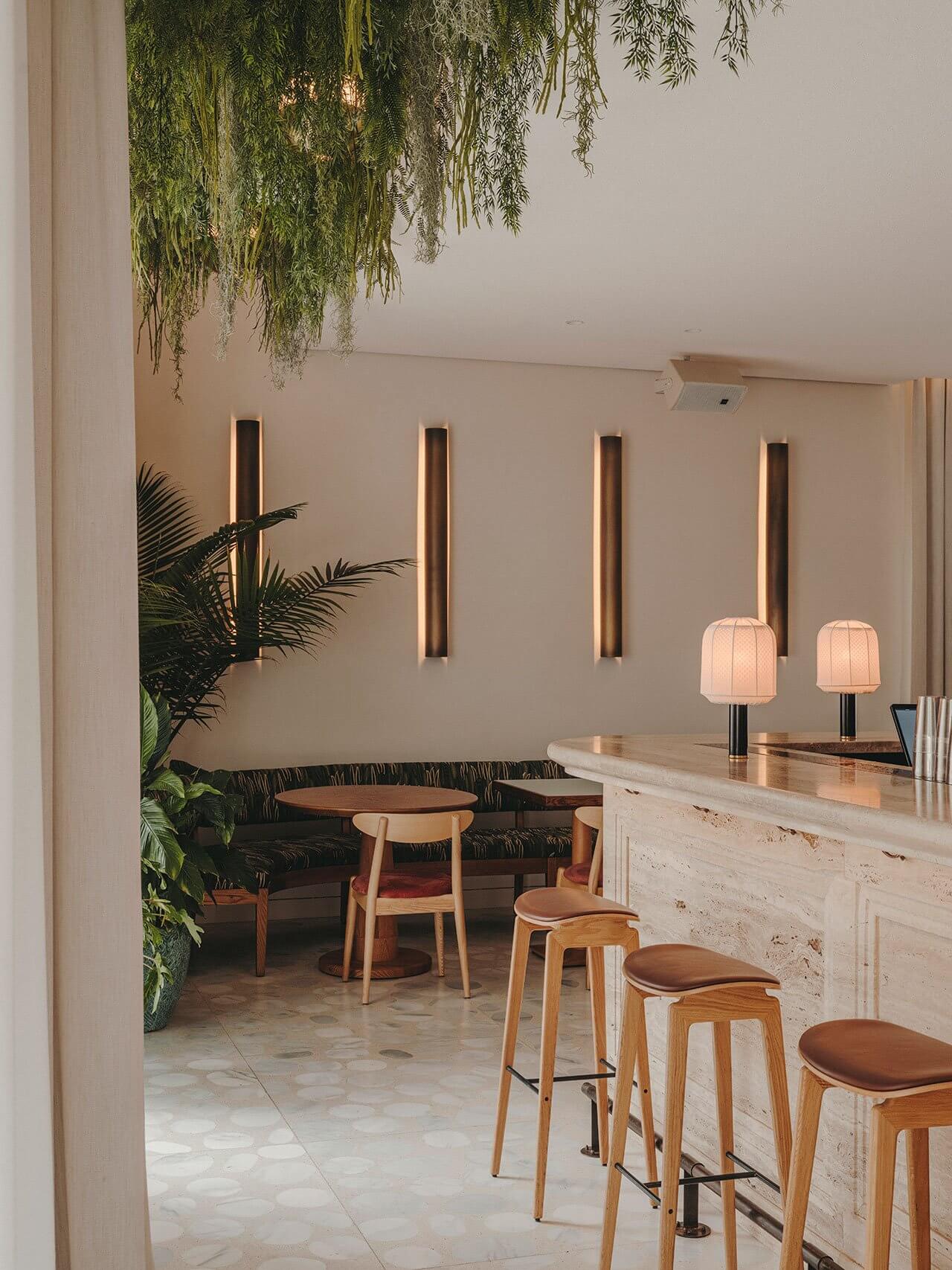 The Standard Ibiza, Balearic Islands, Spain — Modern hospitality interior with travertine bar, wooden stools, suspended greenery, linear wall sconces, and lounge seating in neutral palette.