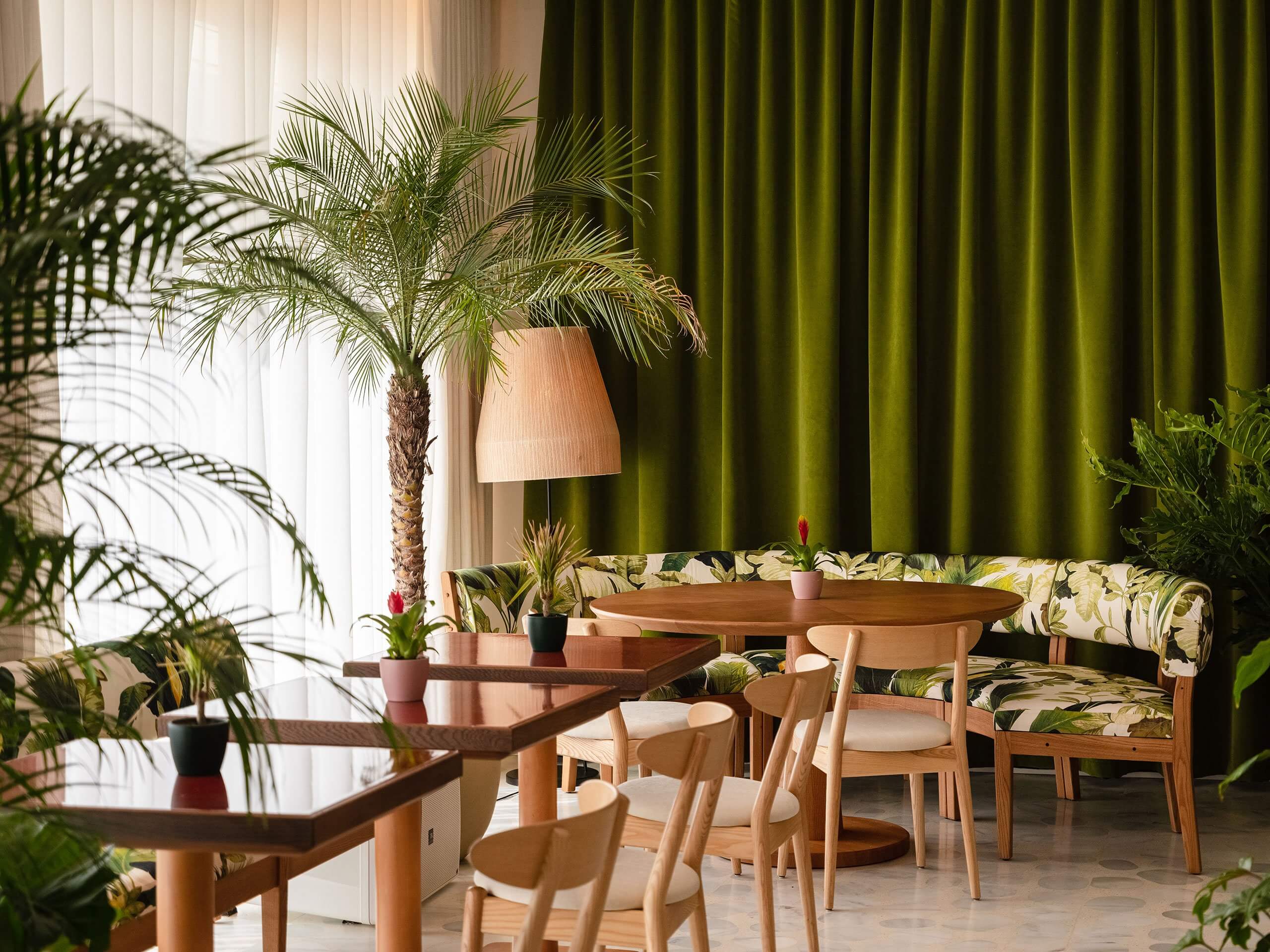 Jara, Balearic Islands — Botanical lounge with mid-century wooden furniture, leafy plants, olive curtains, natural light, and tropical botanical upholstery.