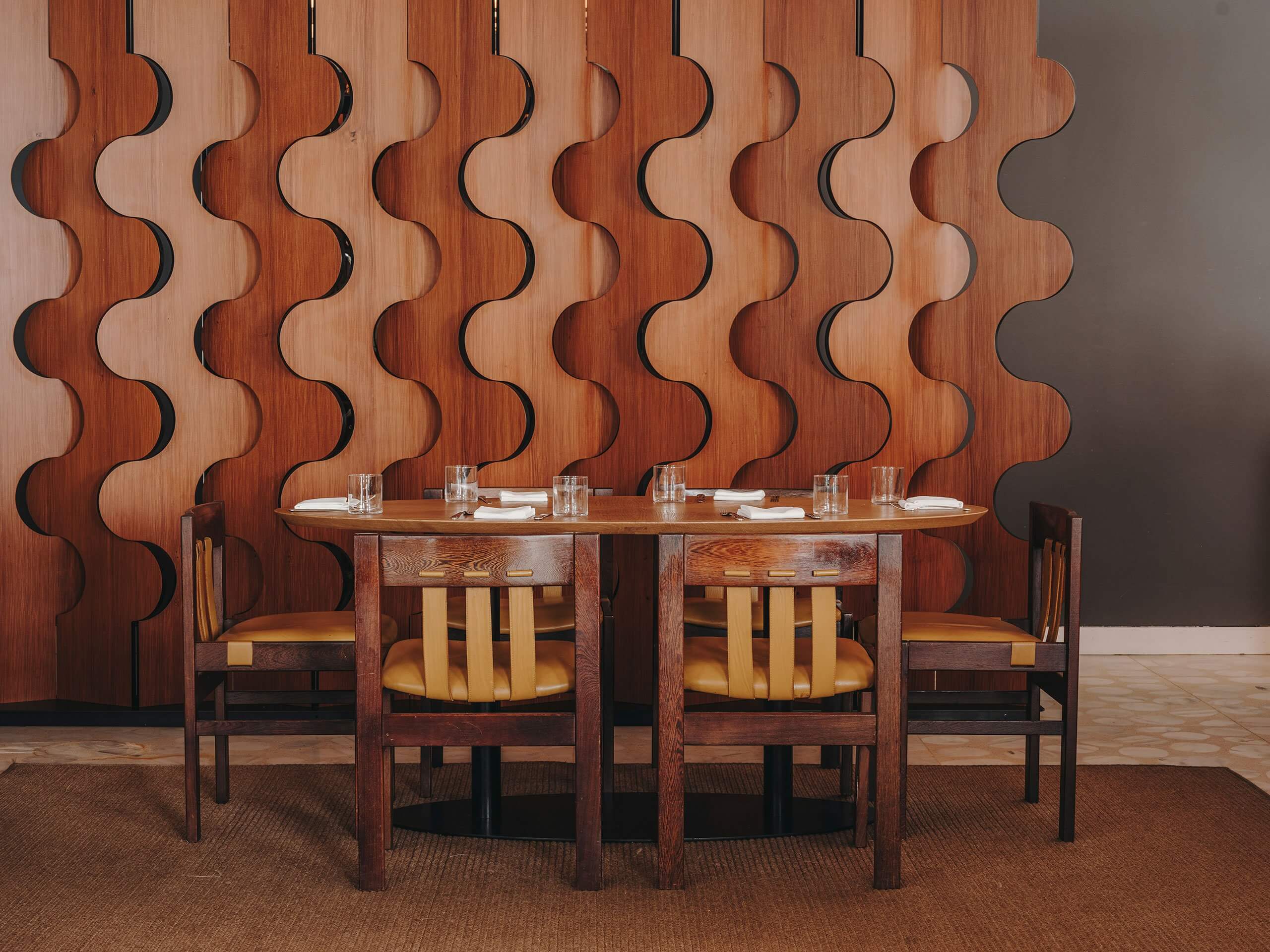 The Standard Ibiza, Balearic Islands, Spain — Dining room with sculptural wooden wall paneling, oval table, ochre leather chairs, minimalist tableware setup.