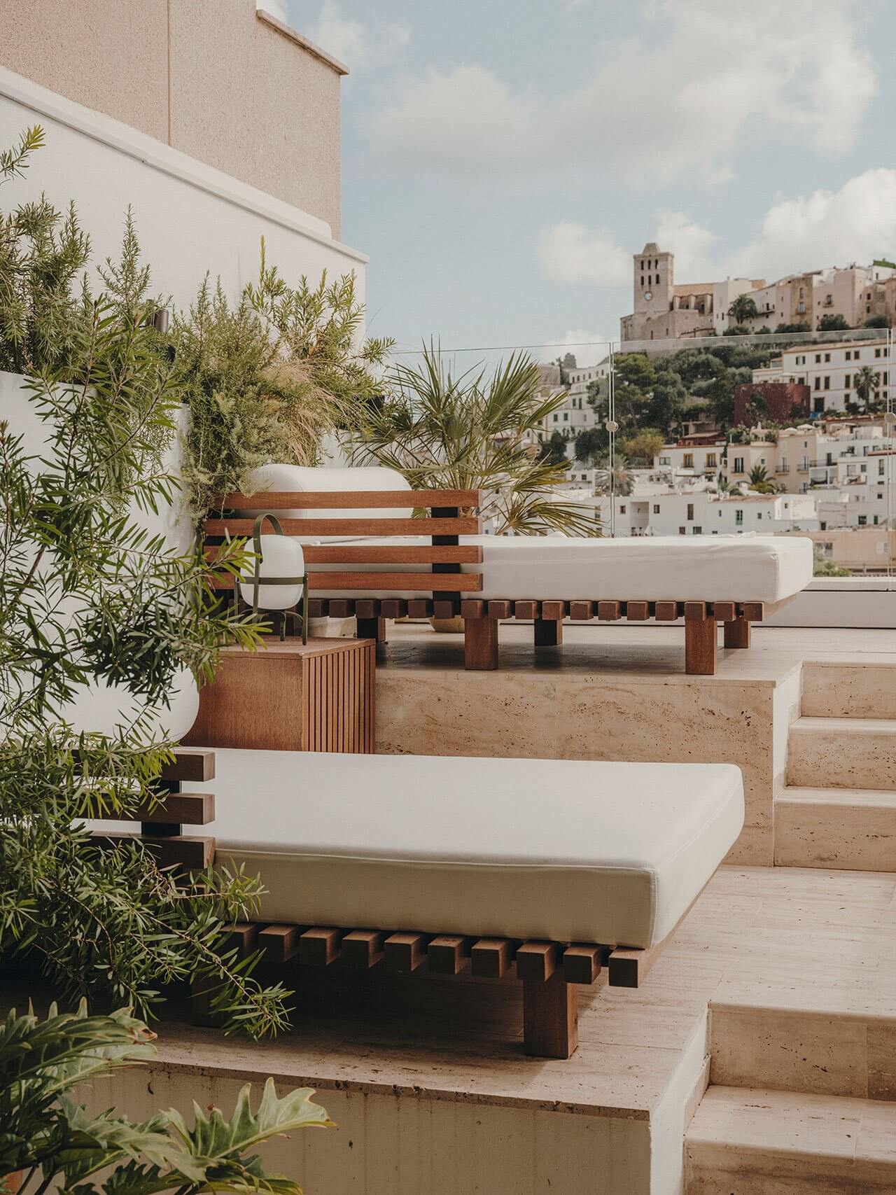 UP, Balearic Islands, Spain — Minimalist rooftop terrace with wooden-slatted daybeds, potted olive trees, and whitewashed architecture overlooking Mediterranean hillside town.