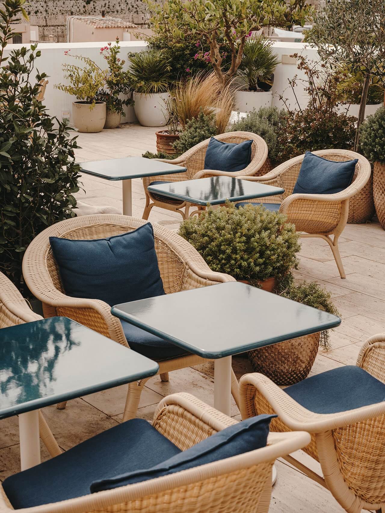 The Standard Ibiza, Balearic Islands, Spain — Natural wicker lounge chairs with navy cushions arranged around pale concrete tables on a sunlit terrace, surrounded by potted plants and white-walled architecture.