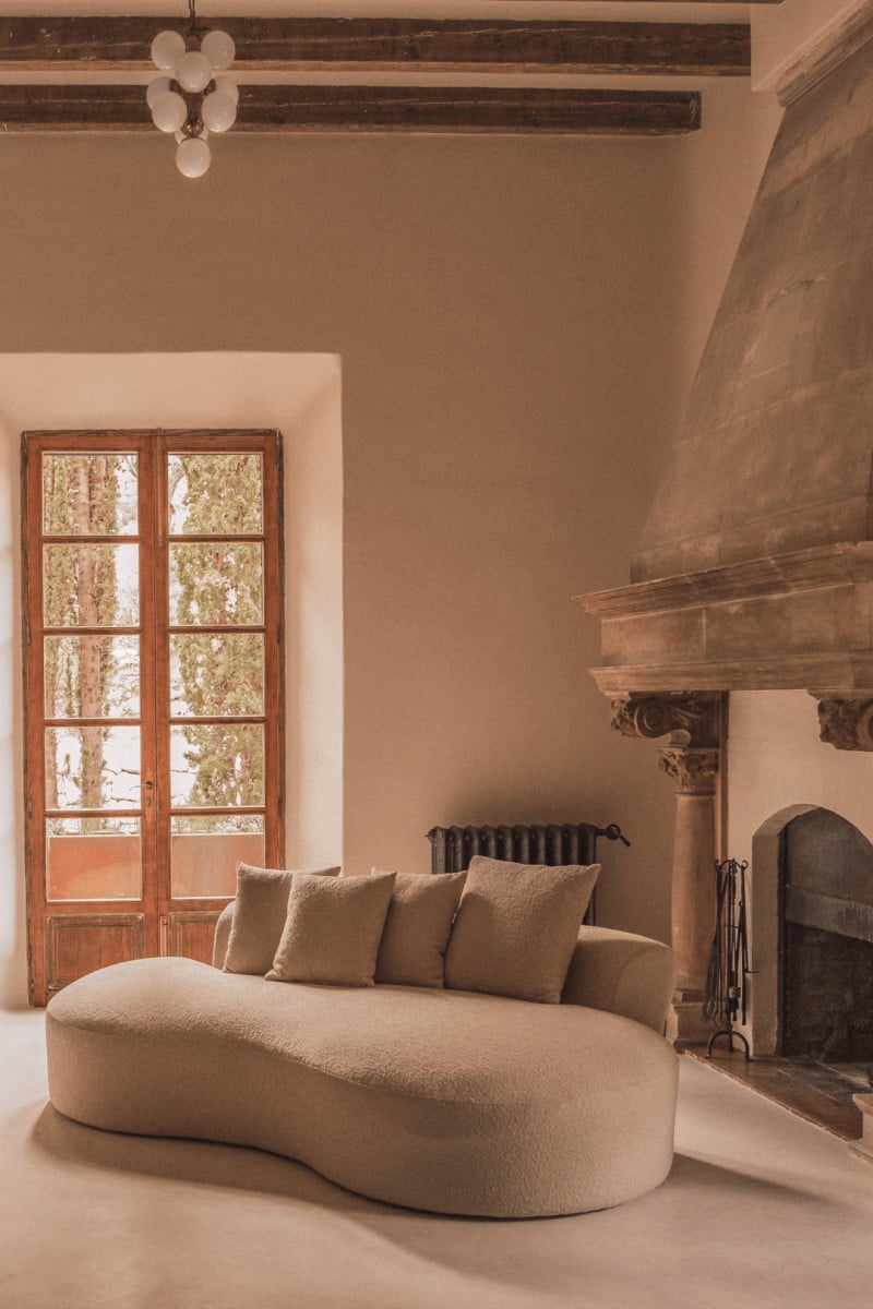 Hotel Corazón, Balearic Islands — Curved cream boucle sofa with pillows before fireplace, wooden beams, arched windows, and minimalist pendant light in warm terracotta interior.