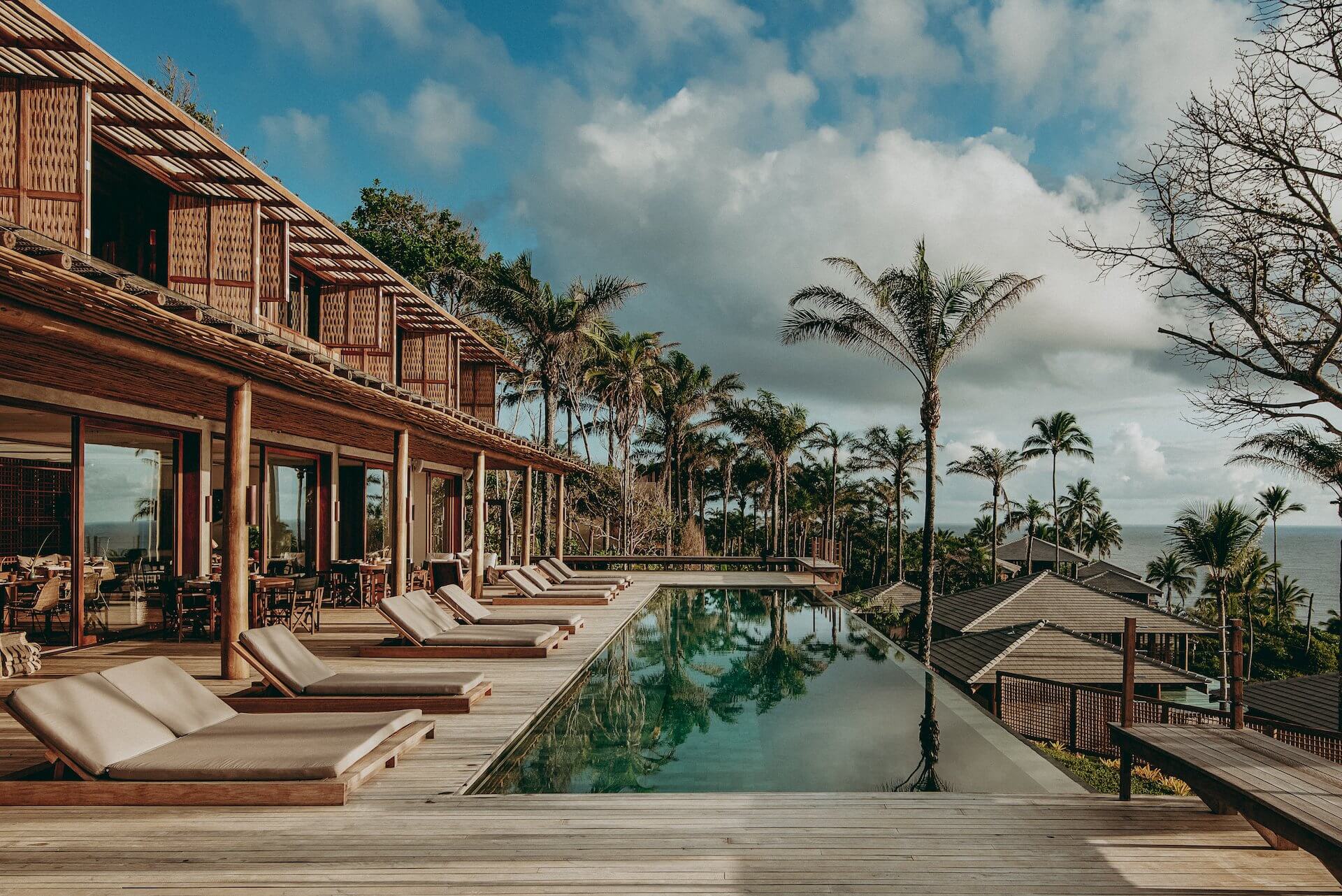 Barracuda Hotel & Villas, South America — Infinity pool with teak loungers, palm trees, and ocean horizon, modern tropical architecture with wooden deck and open-air pavilion.